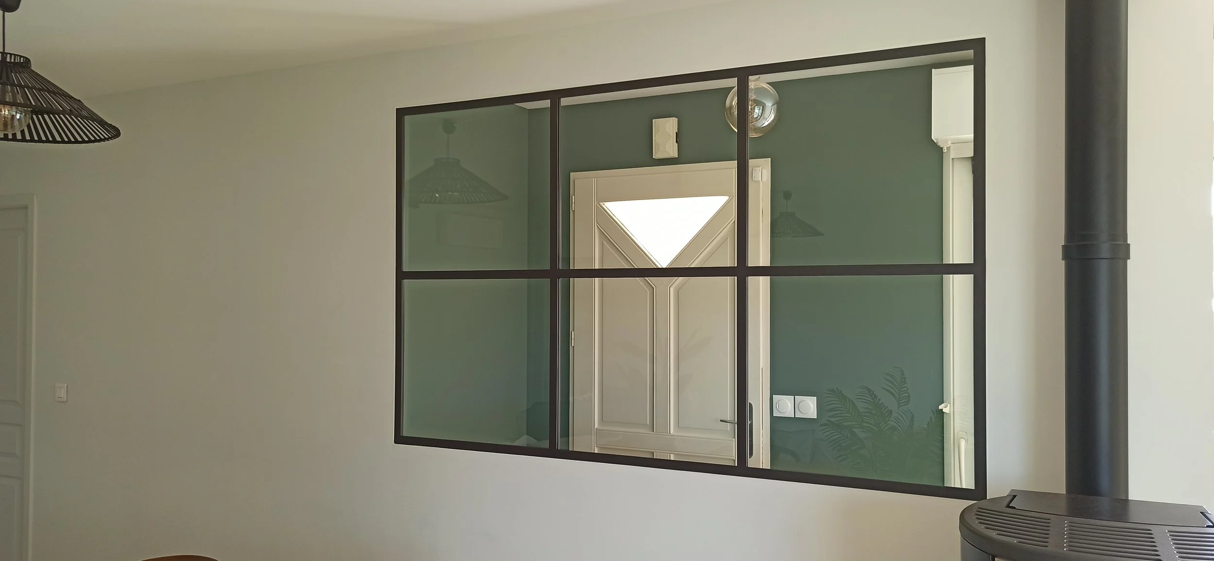 Intérieur d'une maison avec un mur blanc, une fenêtre en verre séparant deux pièces, et une cheminée noire.