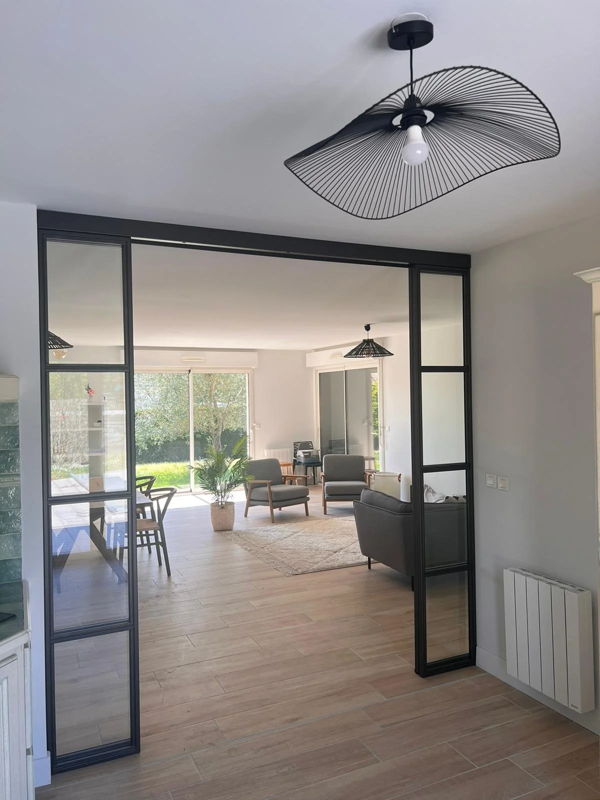 Salon lumineux avec de grandes fenêtres, mobilier moderne avec fauteuils gris et table basse, décor épuré avec une plante verte et un tapis beige.