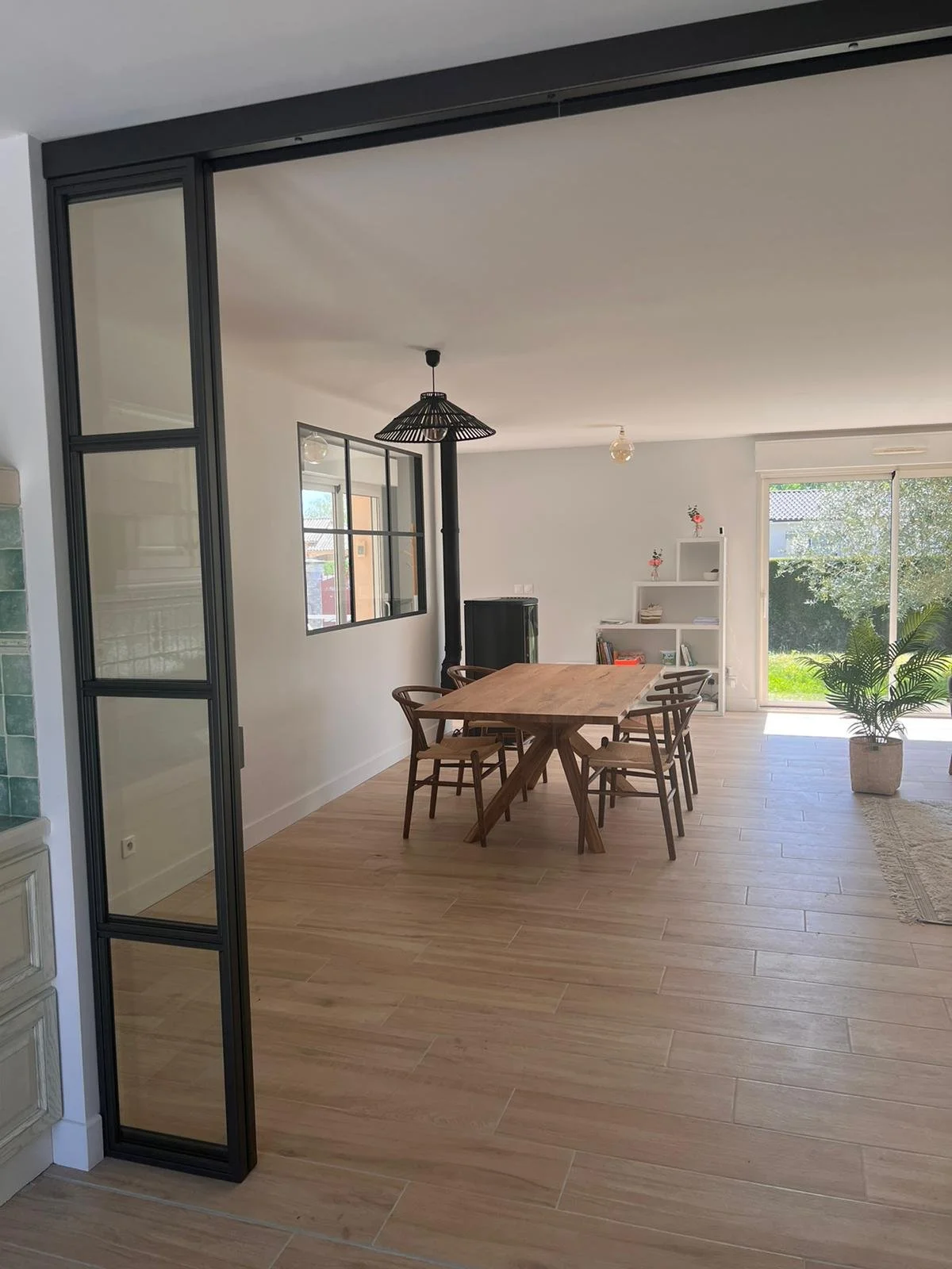 Salle à manger avec une table en bois, six chaises en bois, un luminaire suspendu en rotin, une étagère blanche avec des décorations, un grand fenêtre laissant entrer la lumière naturelle, et une plante d'intérieur dans un pot en rotin.