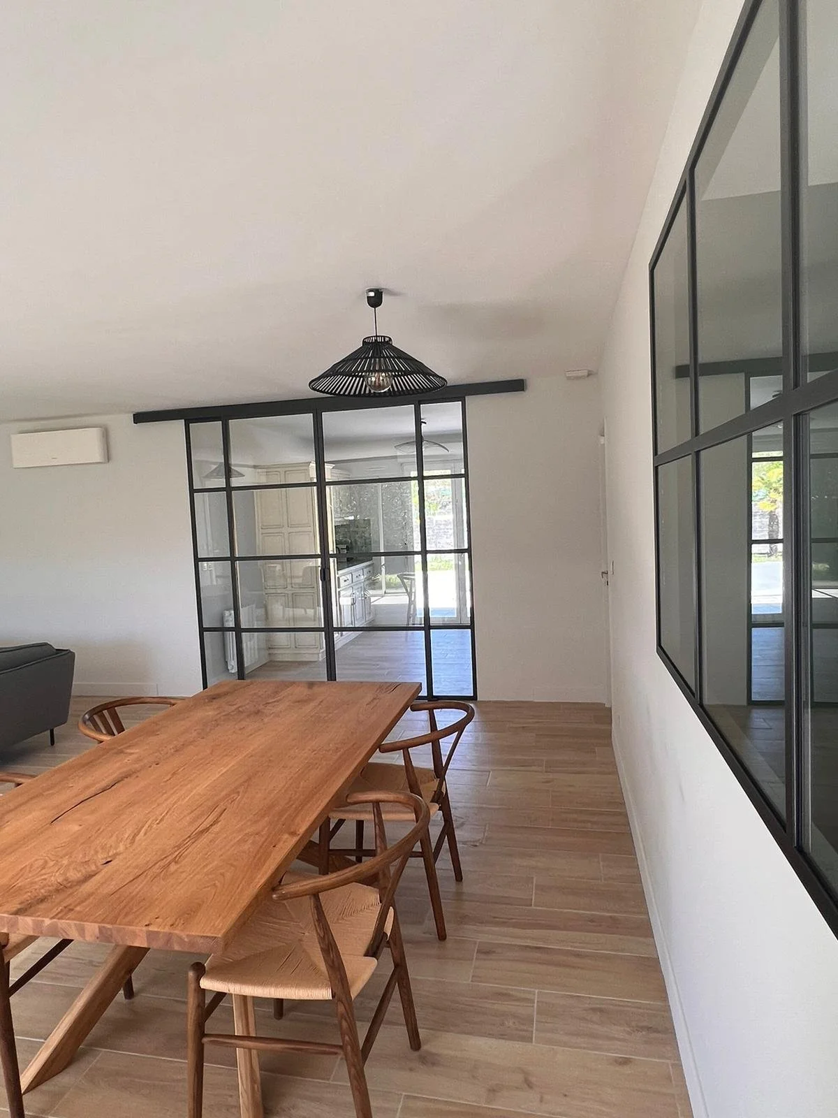 Une salle à manger moderne avec une grande table en bois et des chaises en bois organique. Il y a un luminaire noir suspendu au plafond, et un mur vitré séparant la salle à manger de la cuisine.