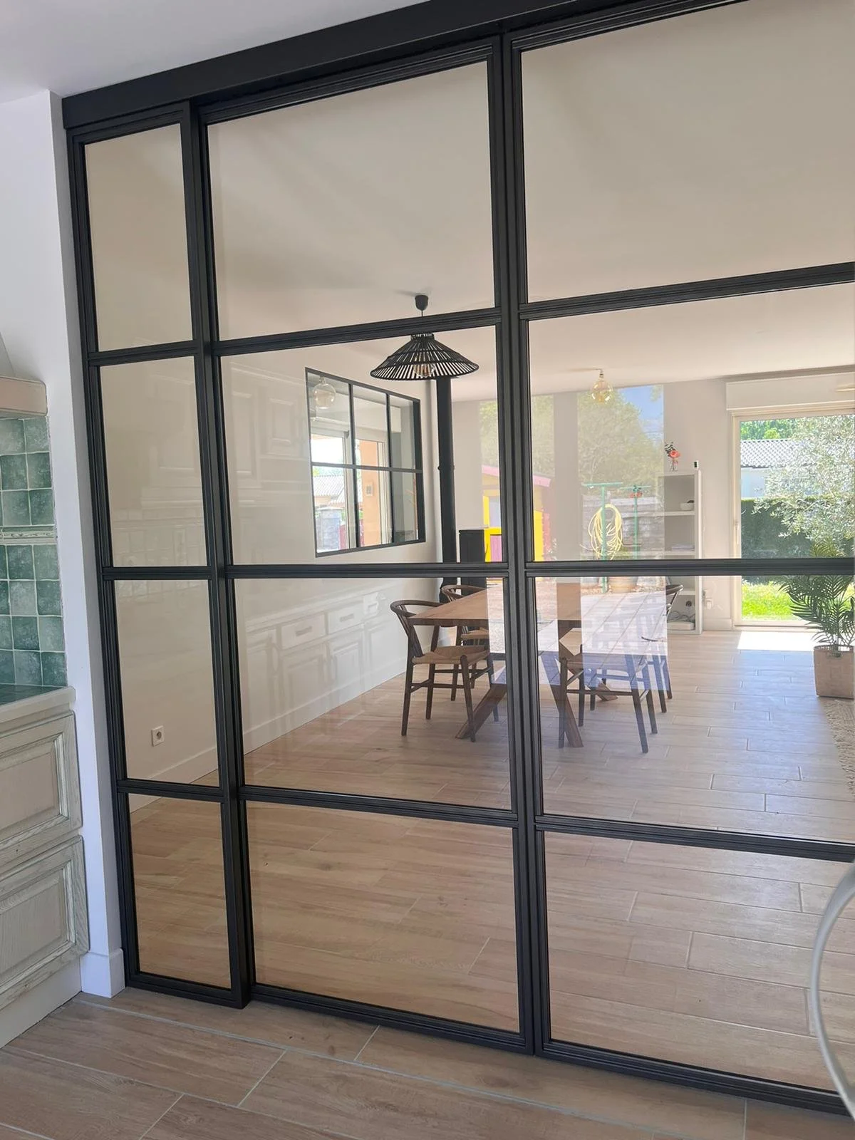 Vue à travers une porte vitrée style fenêtre intérieure, montrant une salle à manger avec une table en bois et six chaises en bois, un plafond blanc avec un luminaire noir, un mur blanc avec un tableau, et une grande fenêtre laissant entrer la lumièr