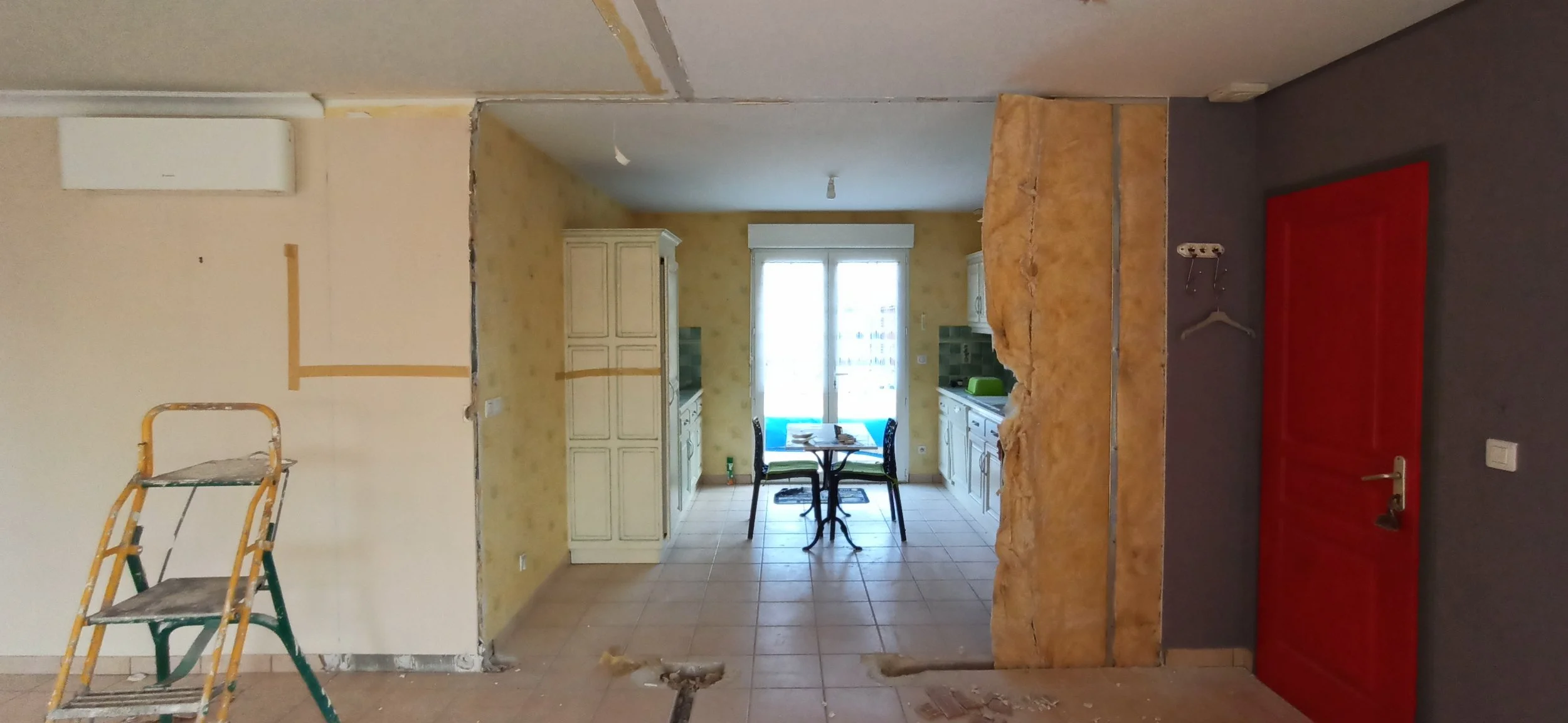 Intérieur d'une cuisine en cours de rénovation avec un mur démoli, une table et des chaises devant une fenêtre, et un mur avec une porte rouge