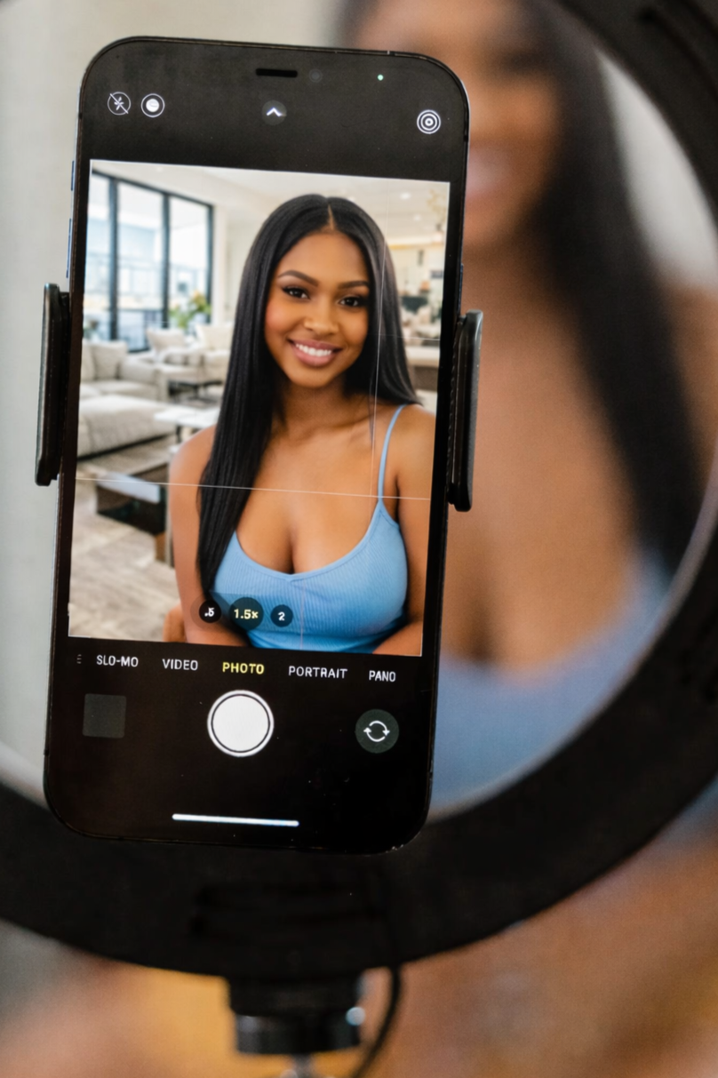 A woman taking a selfie with a smartphone on a tripod inside a bright, modern living room with large windows and white furniture.