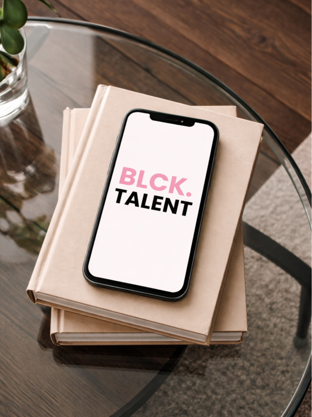 An iPhone displaying the words 'BLACK TALENT' resting on top of two stacked beige books on a glass table, with a plant in a glass container nearby.
