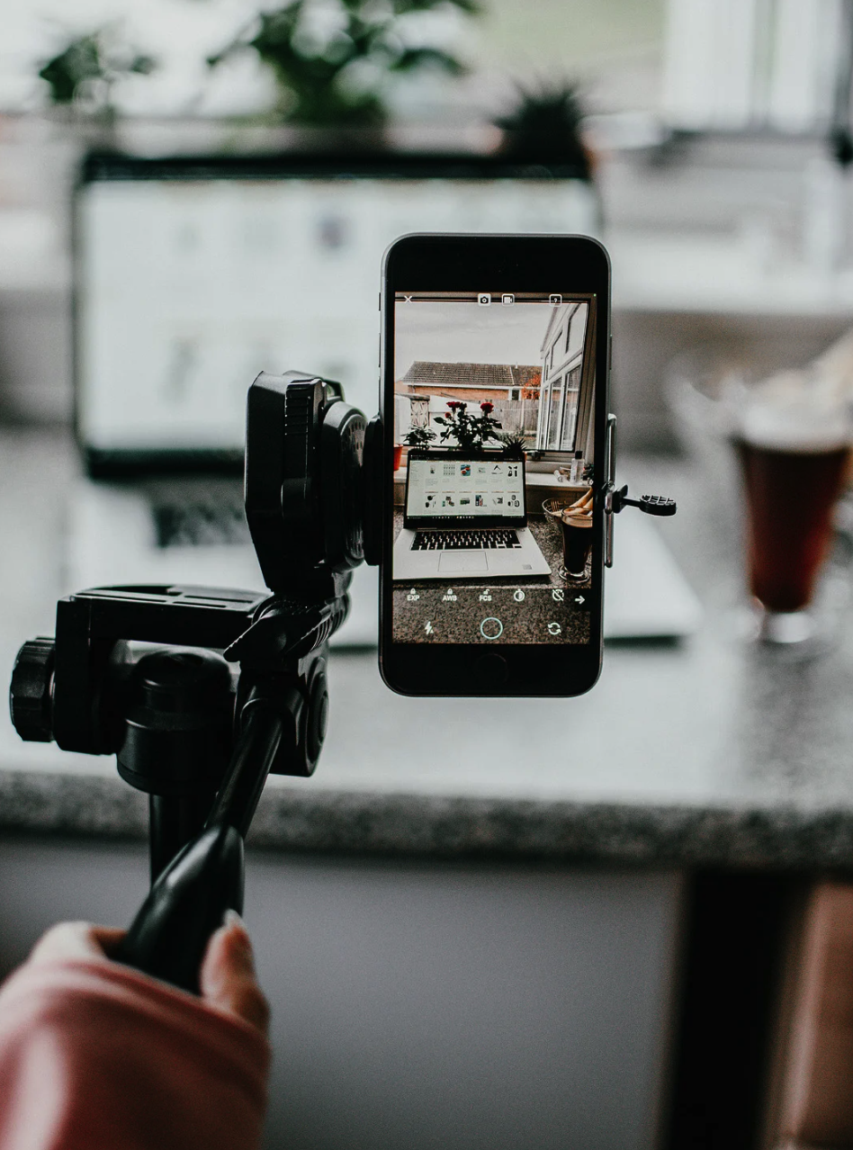 A person using a camera mounted on a tripod to take a photo of a laptop on a kitchen counter, with a window in the background showing a house and plants outside.
