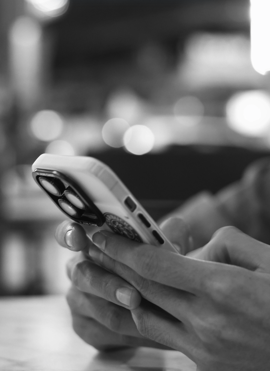 Close-up of a person holding a smartphone with a camera lens, in a blurred indoor setting in black and white.