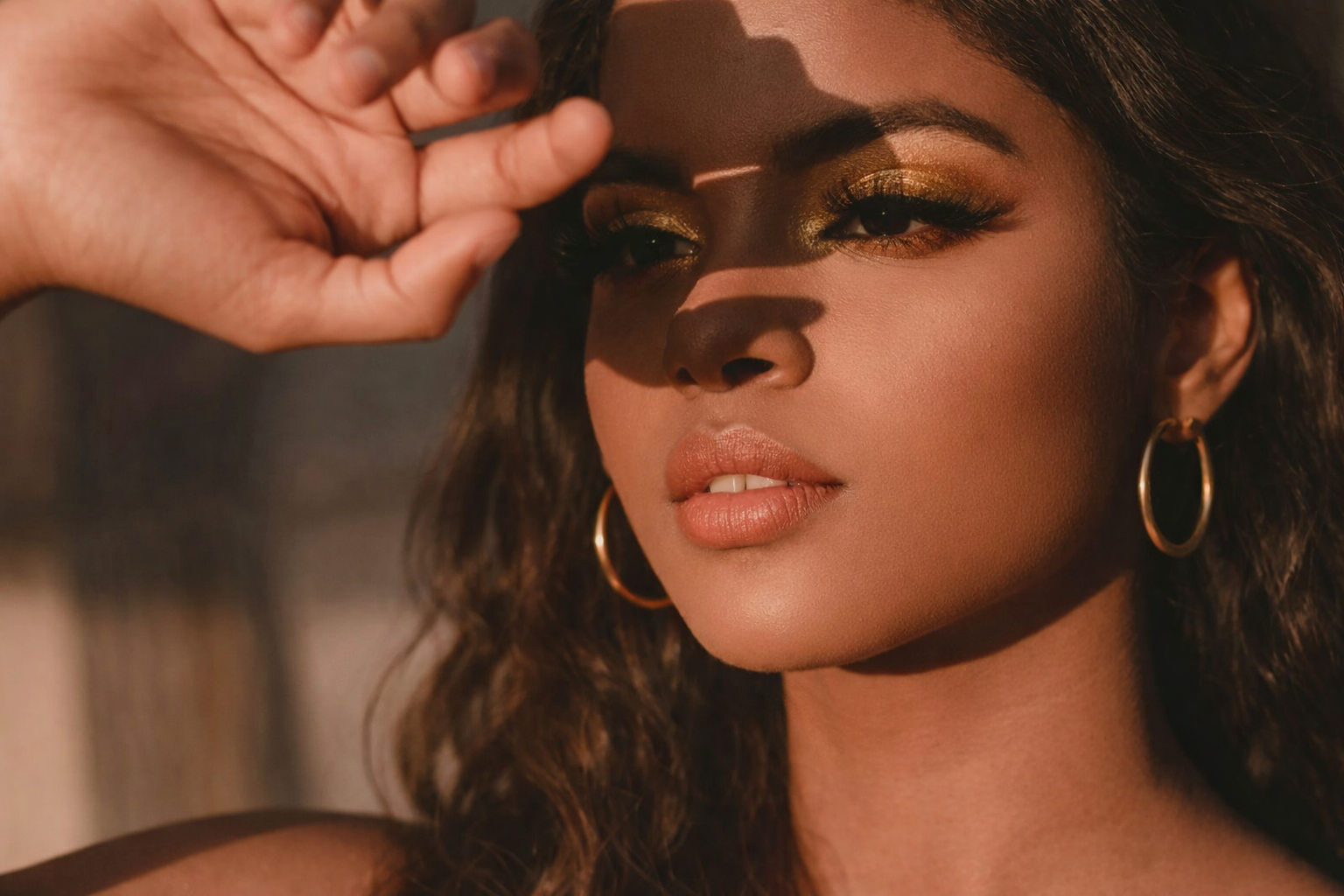 A woman with curly hair, gold eye makeup, and hoop earrings shielding her eyes from the sun with her hand.