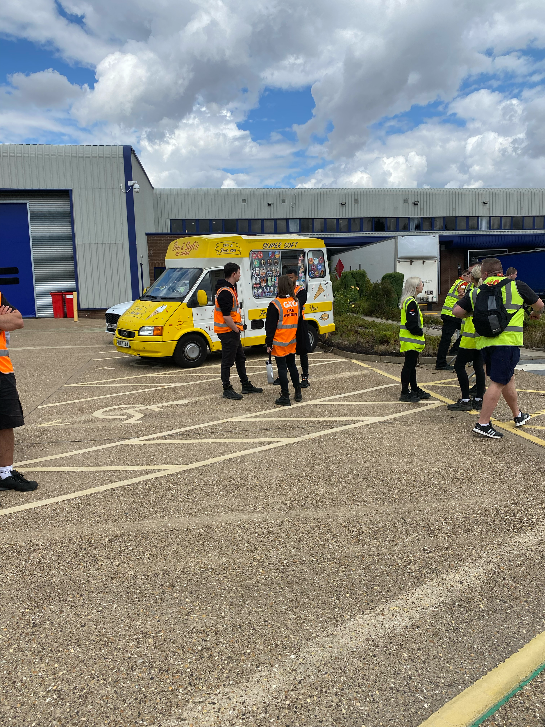 A group of people, some in reflective safety vests, are gathered around an ice cream truck in a parking lot, with a building in the background and partly cloudy sky overhead.