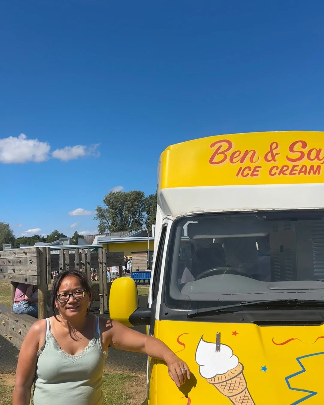 A woman in a light tank top standing next to a yellow ice cream truck with a cartoon ice cream cone illustration and red and yellow text, at an outdoor event on a sunny day.