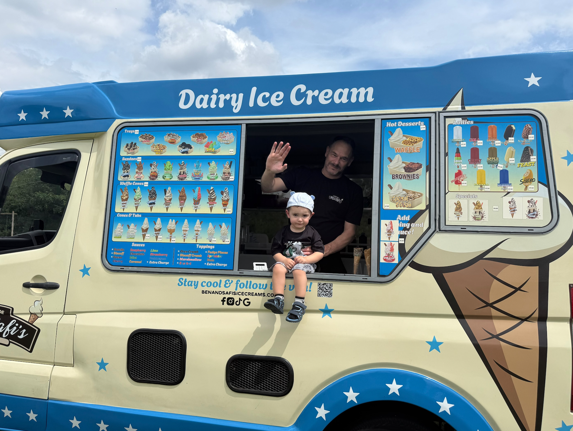 A colorful ice cream truck with large menu displays and a man and a young boy sitting at the window. The truck features various ice cream options, with the words 'Dairy Ice Cream' on top and stars decorating its exterior.