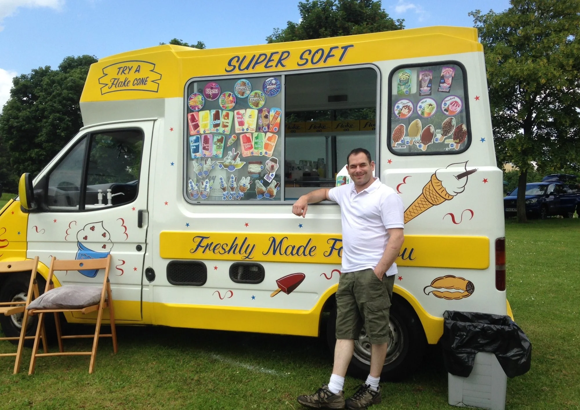 A man standing in front of a colorful ice cream truck on a grassy area. The truck is decorated with ice cream graphics and has a window with various ice cream treats displayed. The man is smiling, wearing a white polo shirt, cargo shorts, and hiking