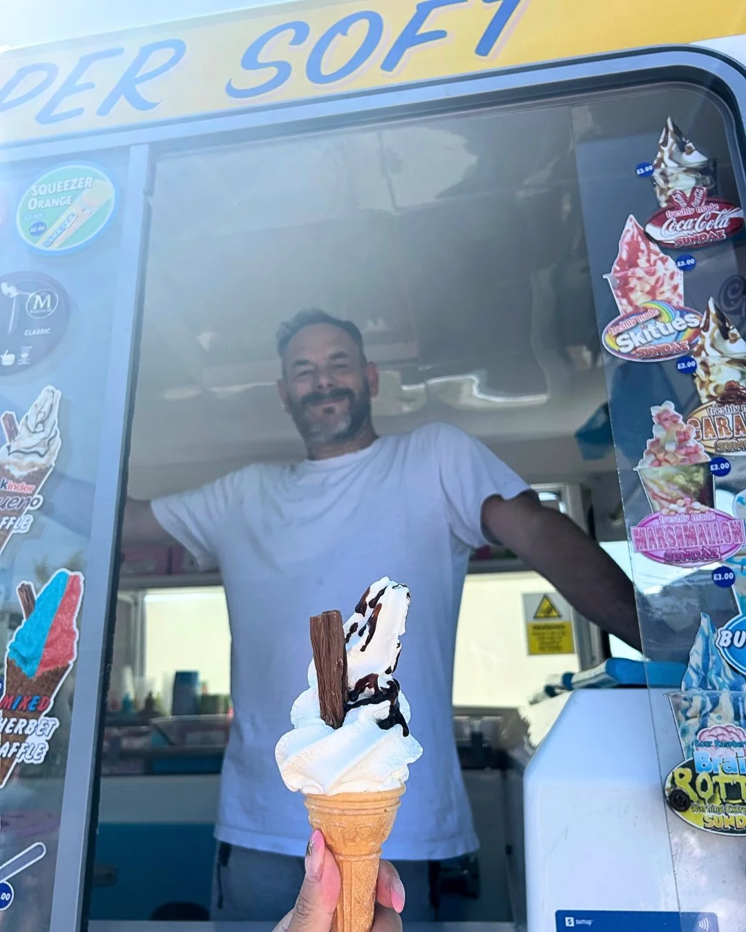 Person holding an ice cream cone with whipped cream, chocolate syrup, and a chocolate wafer, standing behind a glass window at an ice cream stand.