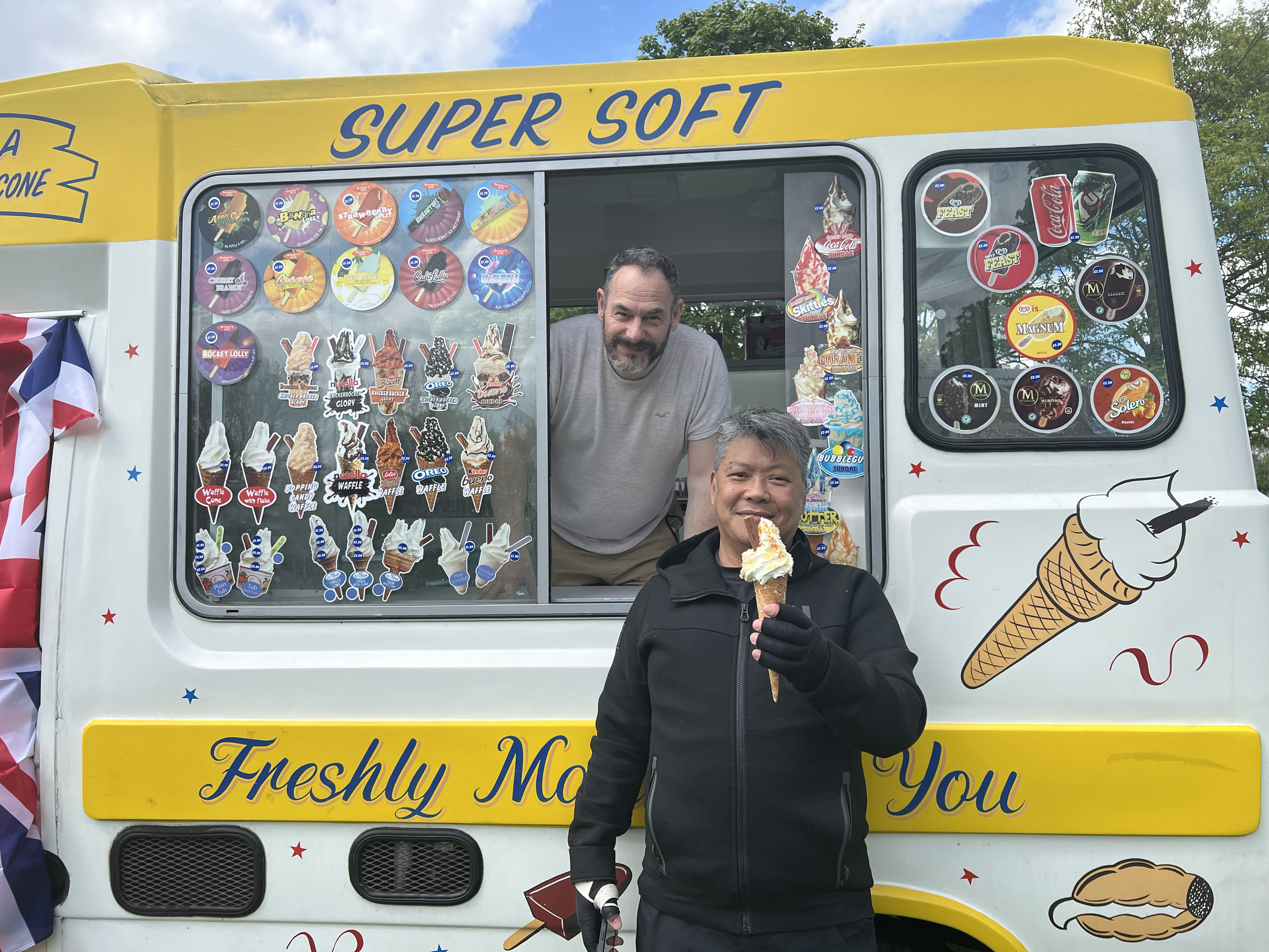 A man holding an ice cream cone standing in front of a colorful ice cream truck with another man inside the truck smiling. The truck has signs and stickers advertising various desserts and brands, with decorative illustrations of ice cream cones.