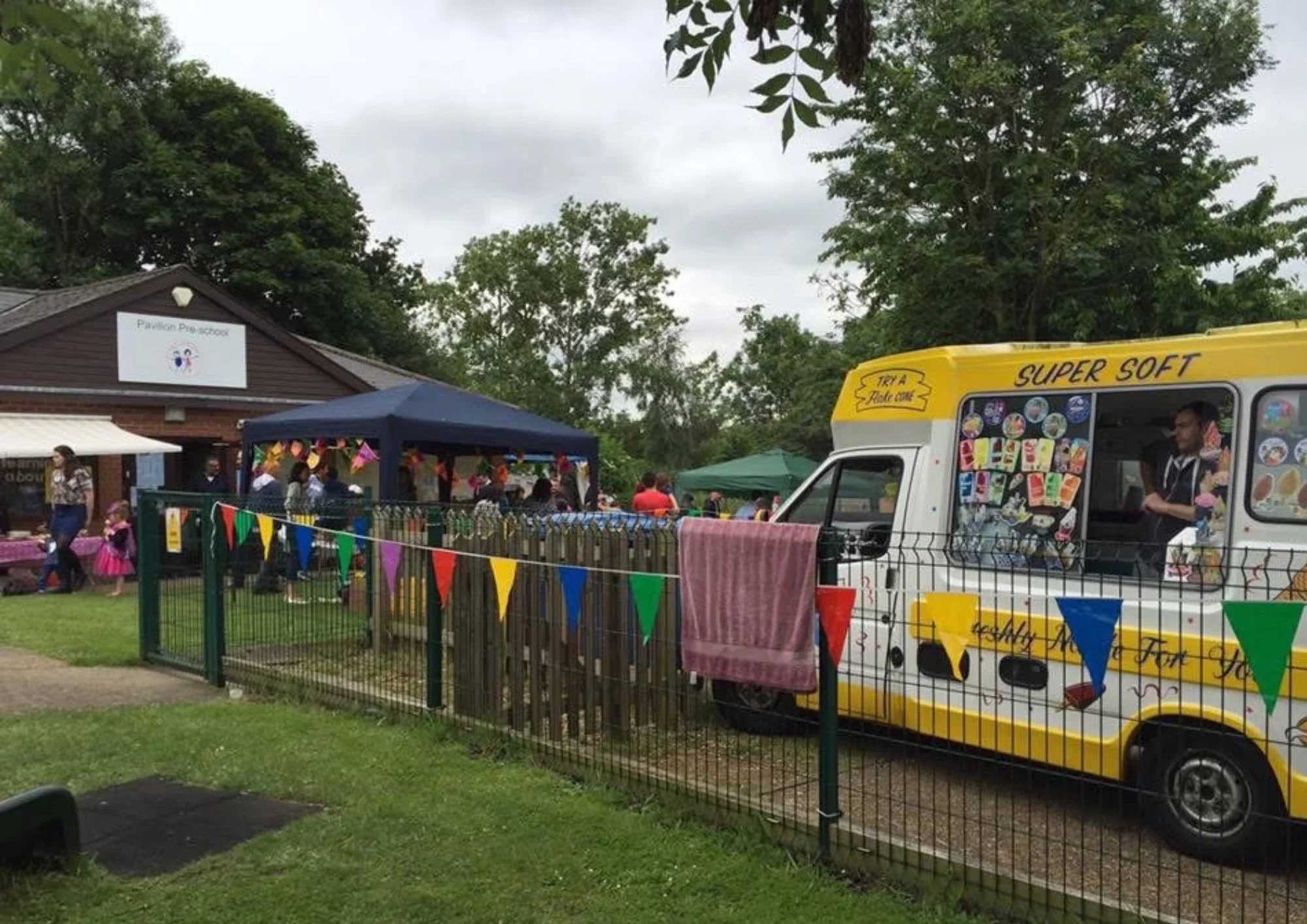 Outdoor community event at a preschool with a yellow ice cream truck, colorful bunting, and a crowd of people gathered near a brown building labeled 'Pavilion Pre-school' on a cloudy day.