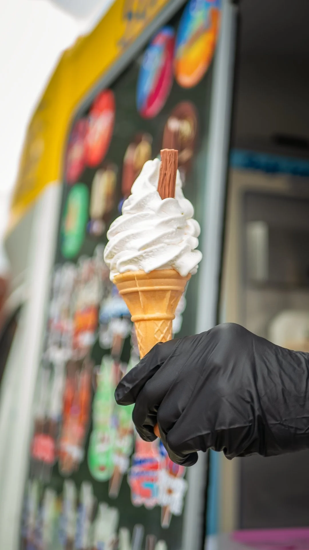 A hand wearing a black glove holding a vanilla soft serve ice cream cone with a chocolate stick on top, in front of a vending machine with colorful ice cream photo displays.