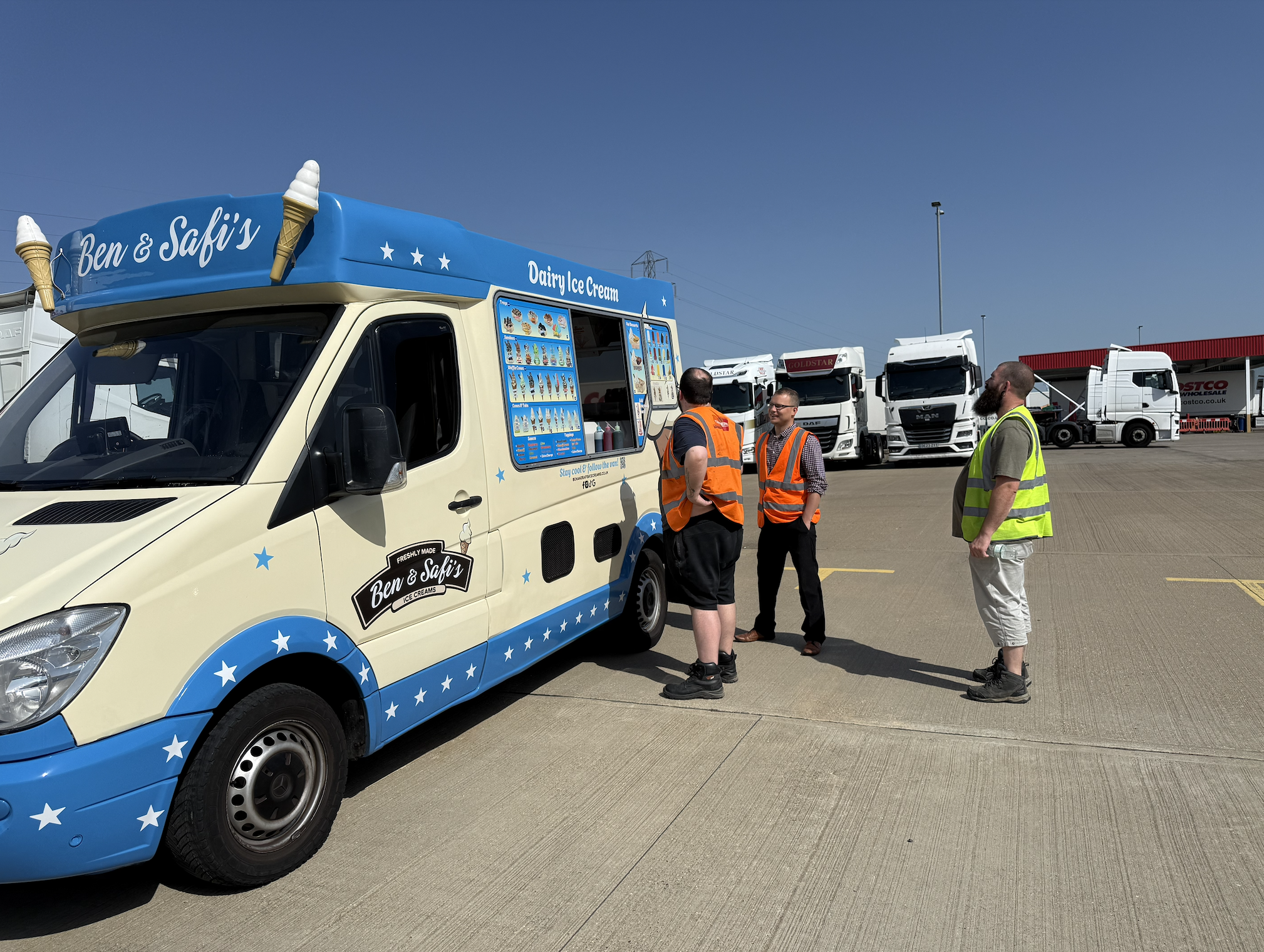 Ice cream truck with Ben & Safi's branding and ice cream cone decorations, parked in an outdoor lot with several trucks and people in safety vests, under a clear blue sky.