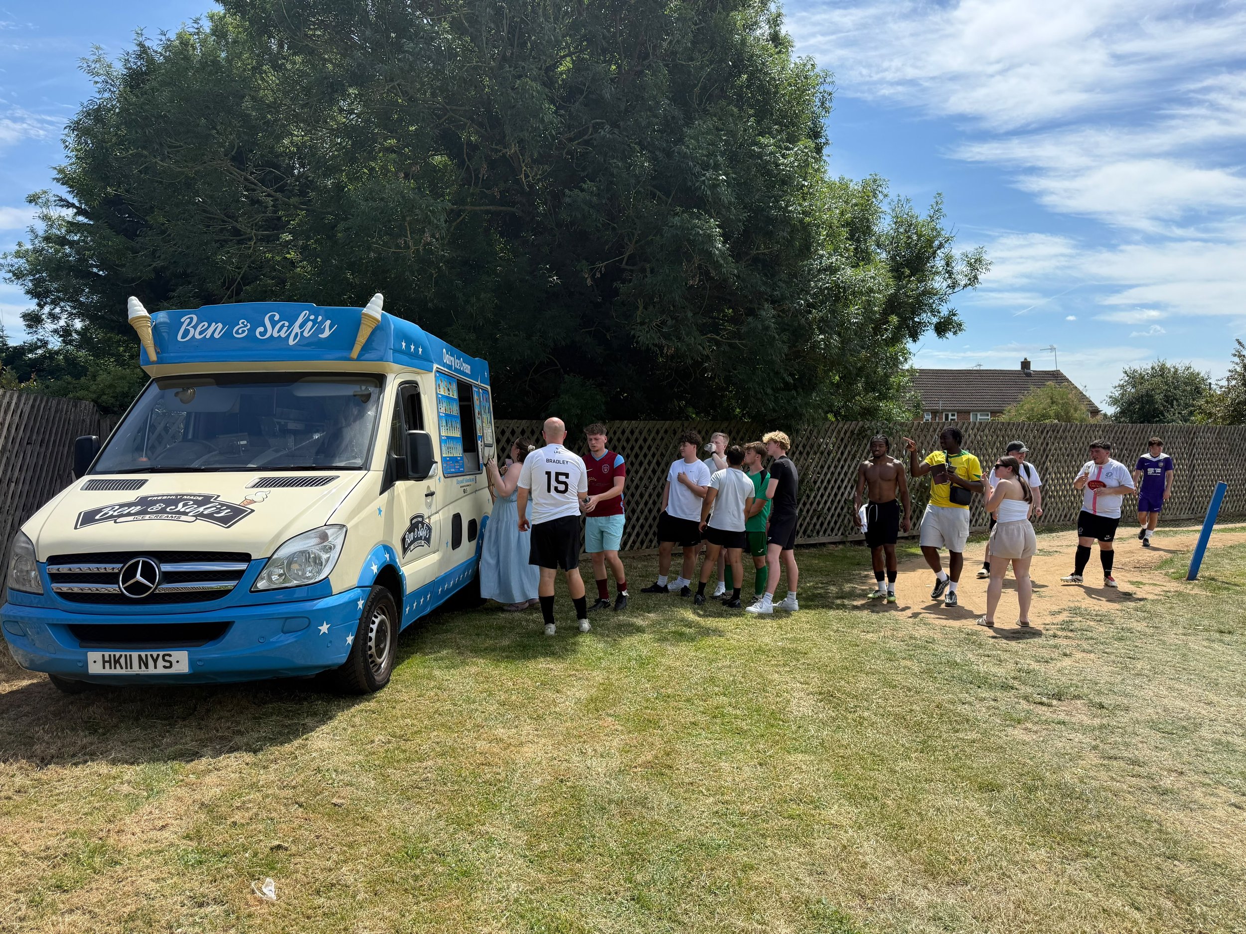A group of people gathered around a Ben & Safi's ice cream truck outdoors on a sunny day, with some people in sports attire and others casually dressed. There is a large tree in the background and a wooden fence behind the group.