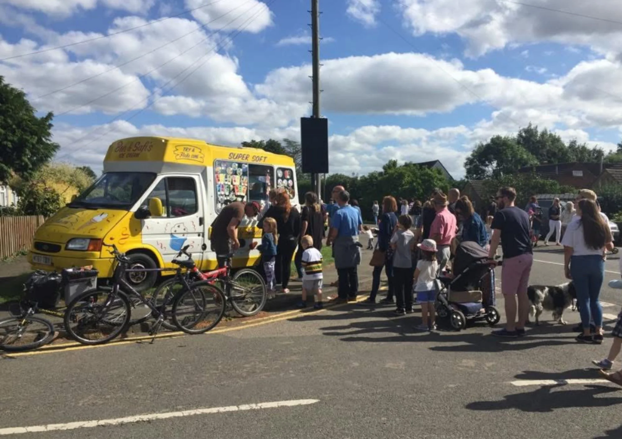 Crowd gathered around a yellow ice cream truck on a sunny day with blue skies and scattered clouds. Several bicycles are parked nearby, and people of various ages are in line or waiting, with some accompanied by dogs. Residential houses are visible i