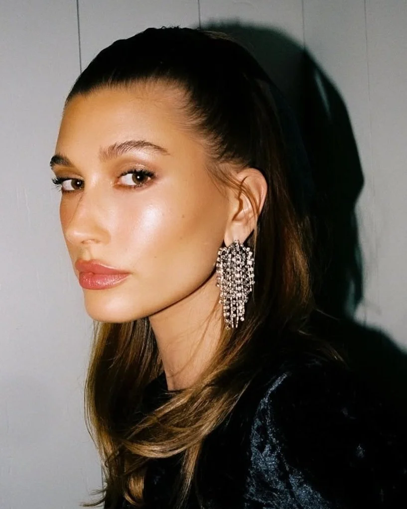 Hailey Bieber, a woman with long, brown hair, wearing large, sparkly chandelier earrings designed by Dannijo and manufactured by Manhattan Rhinestone.