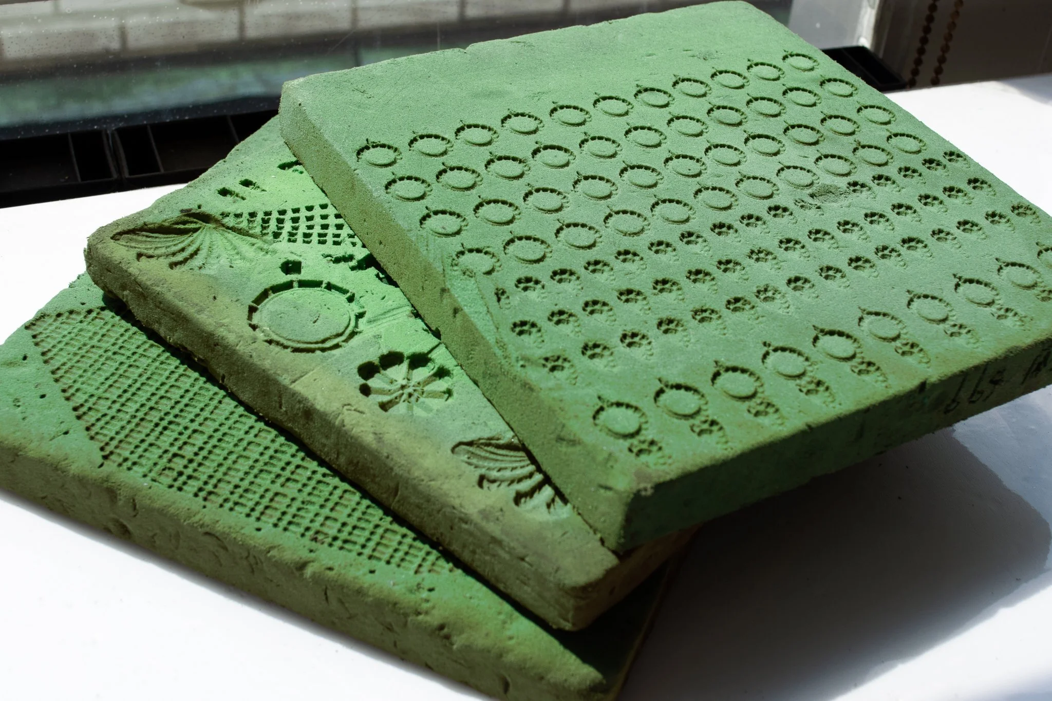 Two green foam printing stamps with intricate patterns, stacked on a white surface near a window.