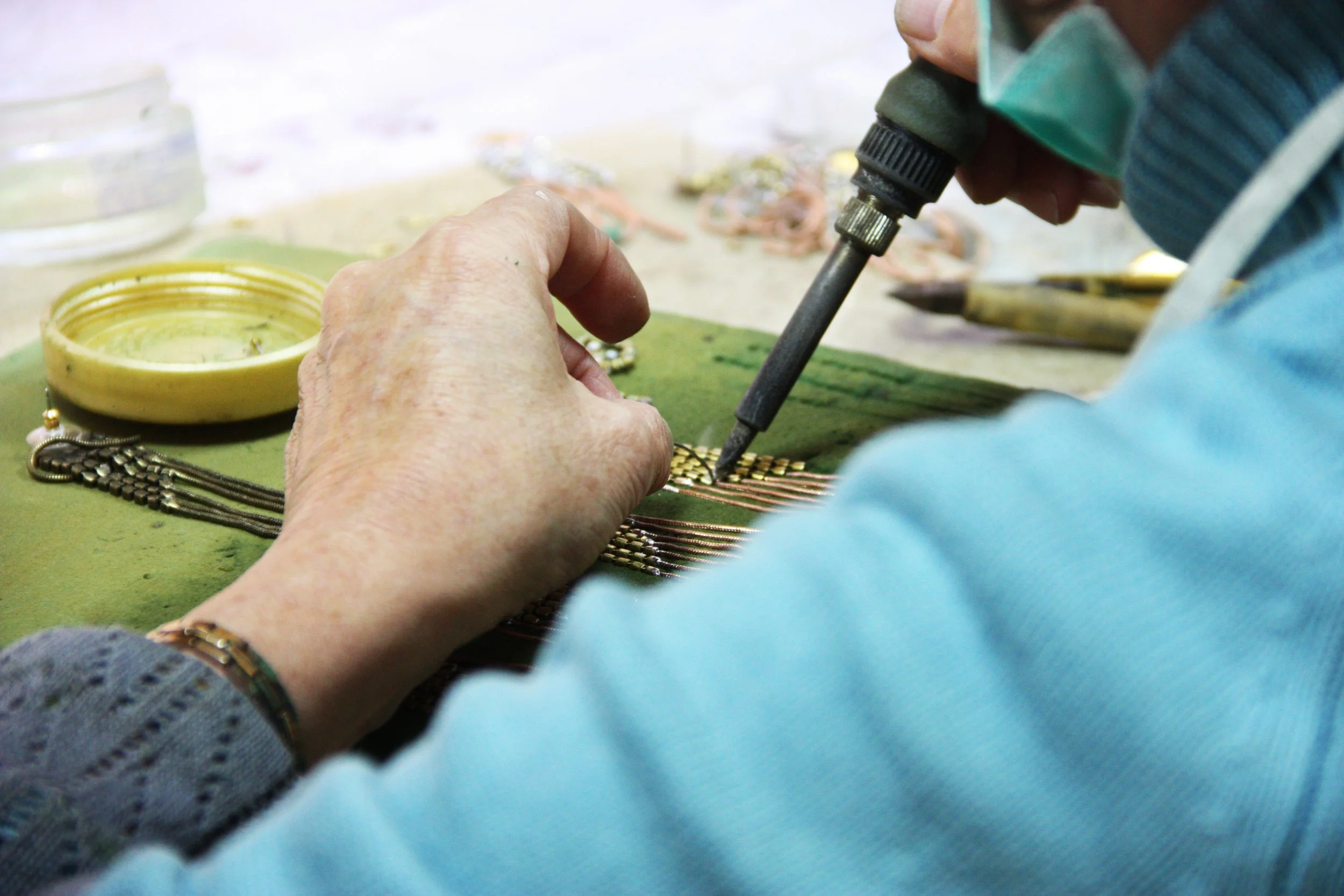A person wearing a face mask and blue sweater soldering or repairing a jewelry piece using a hot soldering iron on a workbench.