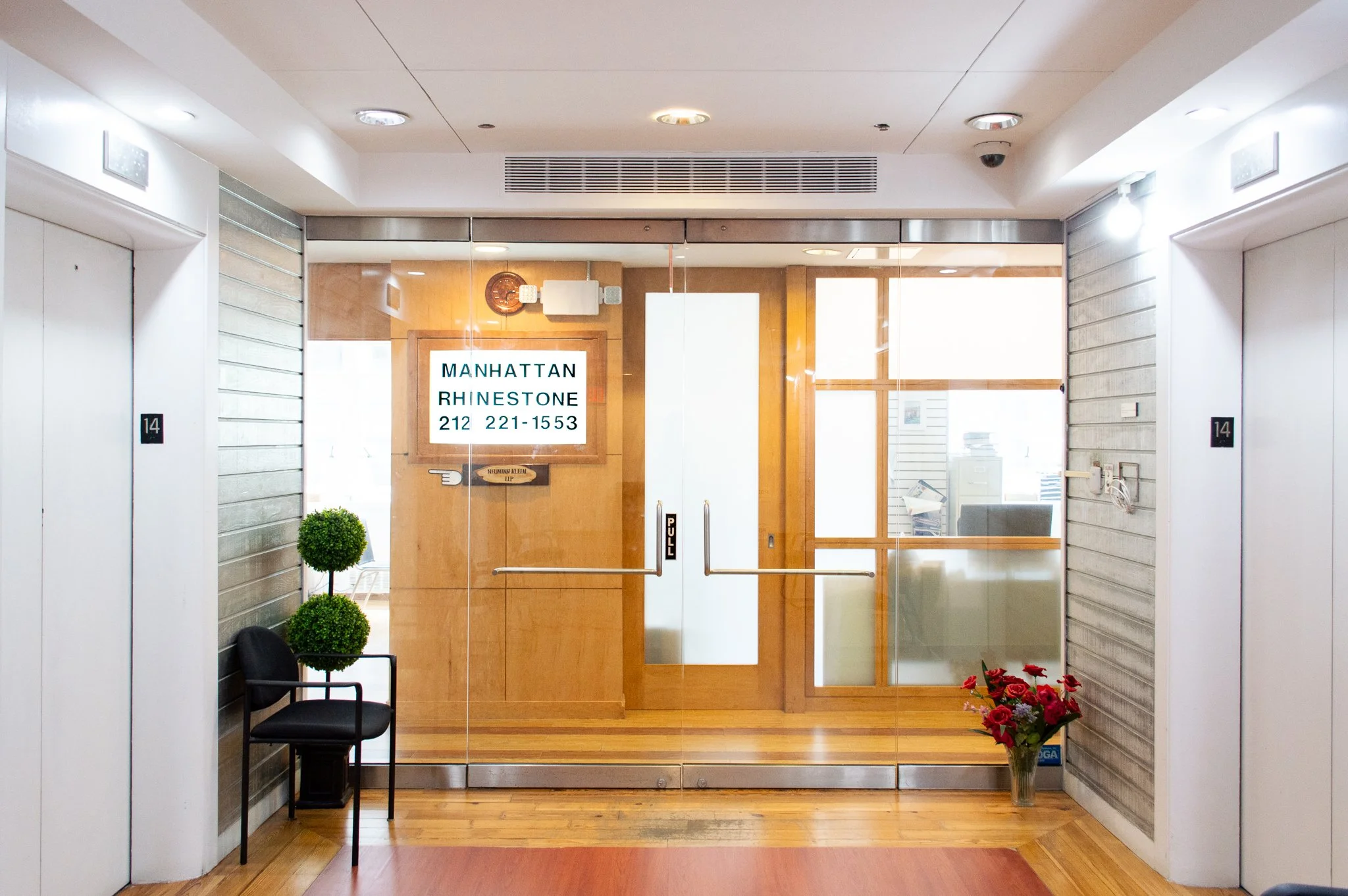 Glass doors leading to an office space with a digital sign displaying Manhattan Rhinestone's custom jewelry manufacturing business information.
