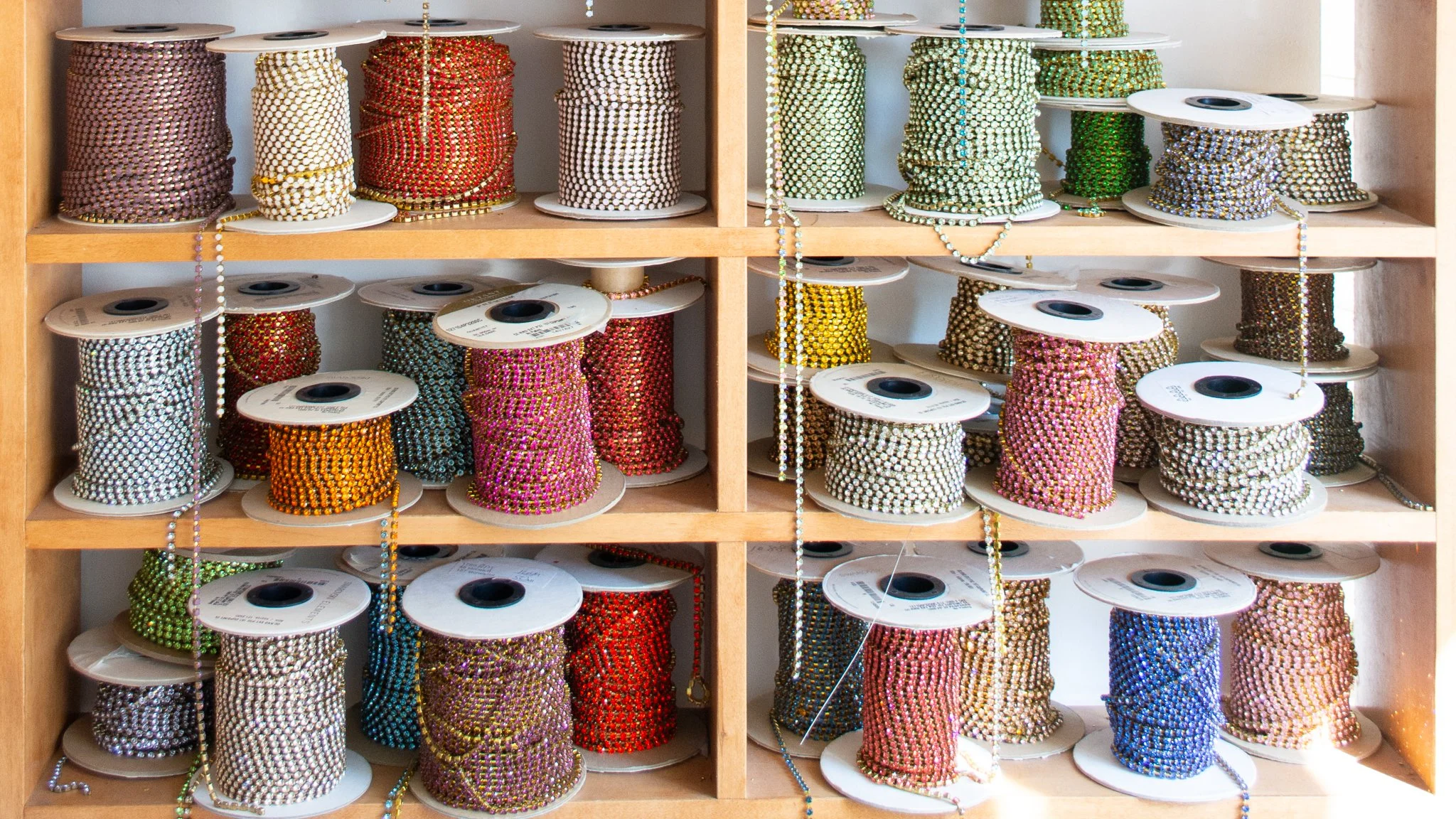 Spools of colorful rhinestone chains on wooden shelves, with some rhinestones draped over the spools.