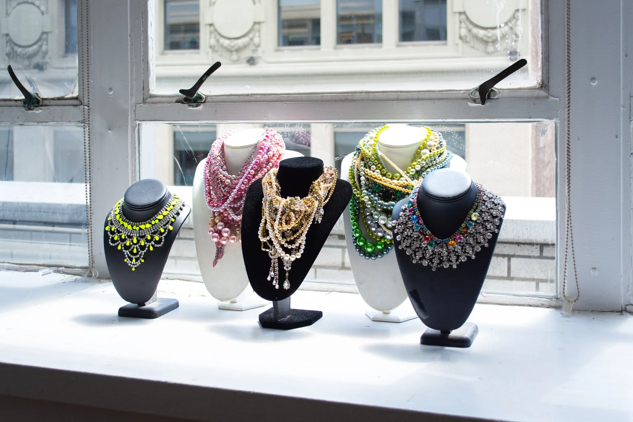 Display of colorful beaded necklaces on black and white busts on a windowsill with an urban building outside.