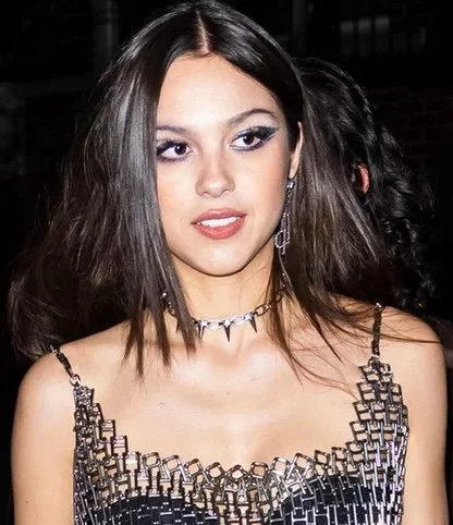 Olivia Rodrigo, a young woman with shoulder-length brown hair, wearing a black and silver patterned dress, and a silver metallic necklace designed by Joomi Lim and manufactured by Manhattan Rhinestone.