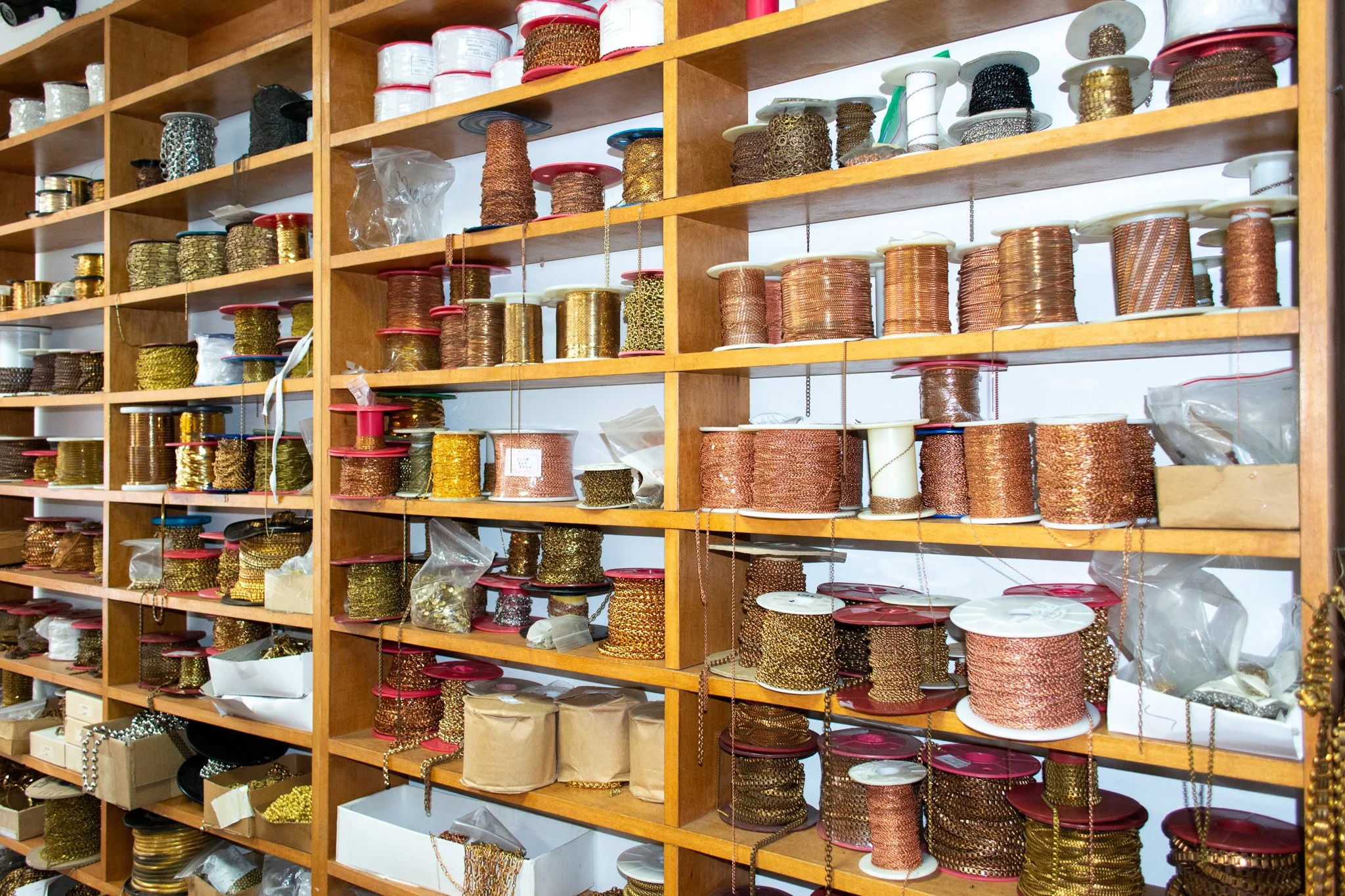 Wooden shelves filled with spools of metallic and decorative chains and cords in various colors including gold, copper, silver, and black, stored in boxes, cups, and plastic bags.