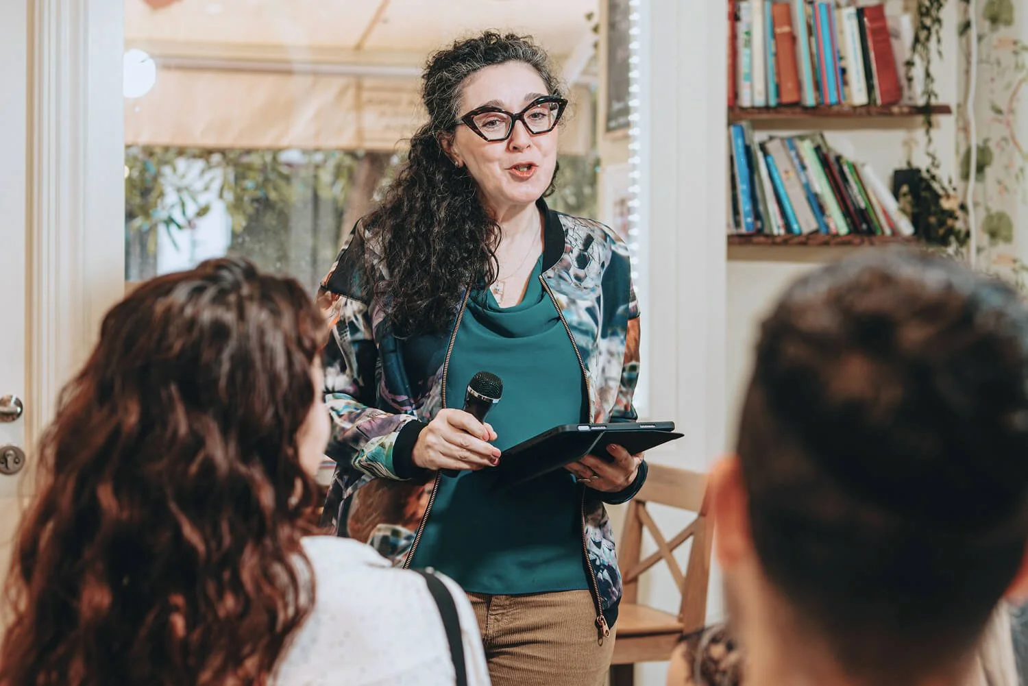 A woman with curly dark hair, glasses, and a patterned jacket holding a microphone and a tablet, speaking to a group of people in a cozy room with a bookshelf in the background.