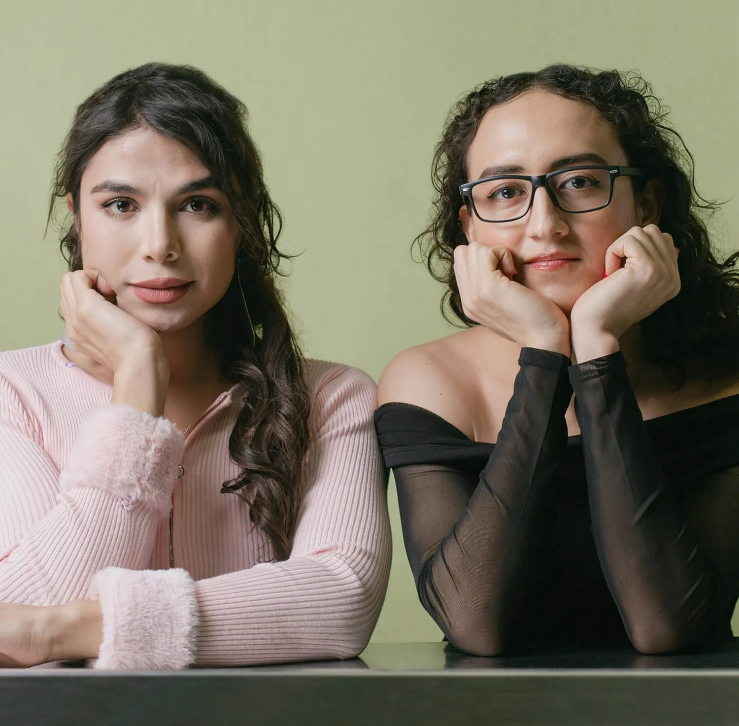 Two women sitting at a table with their heads resting on their hands, looking at the camera against a pale green background.