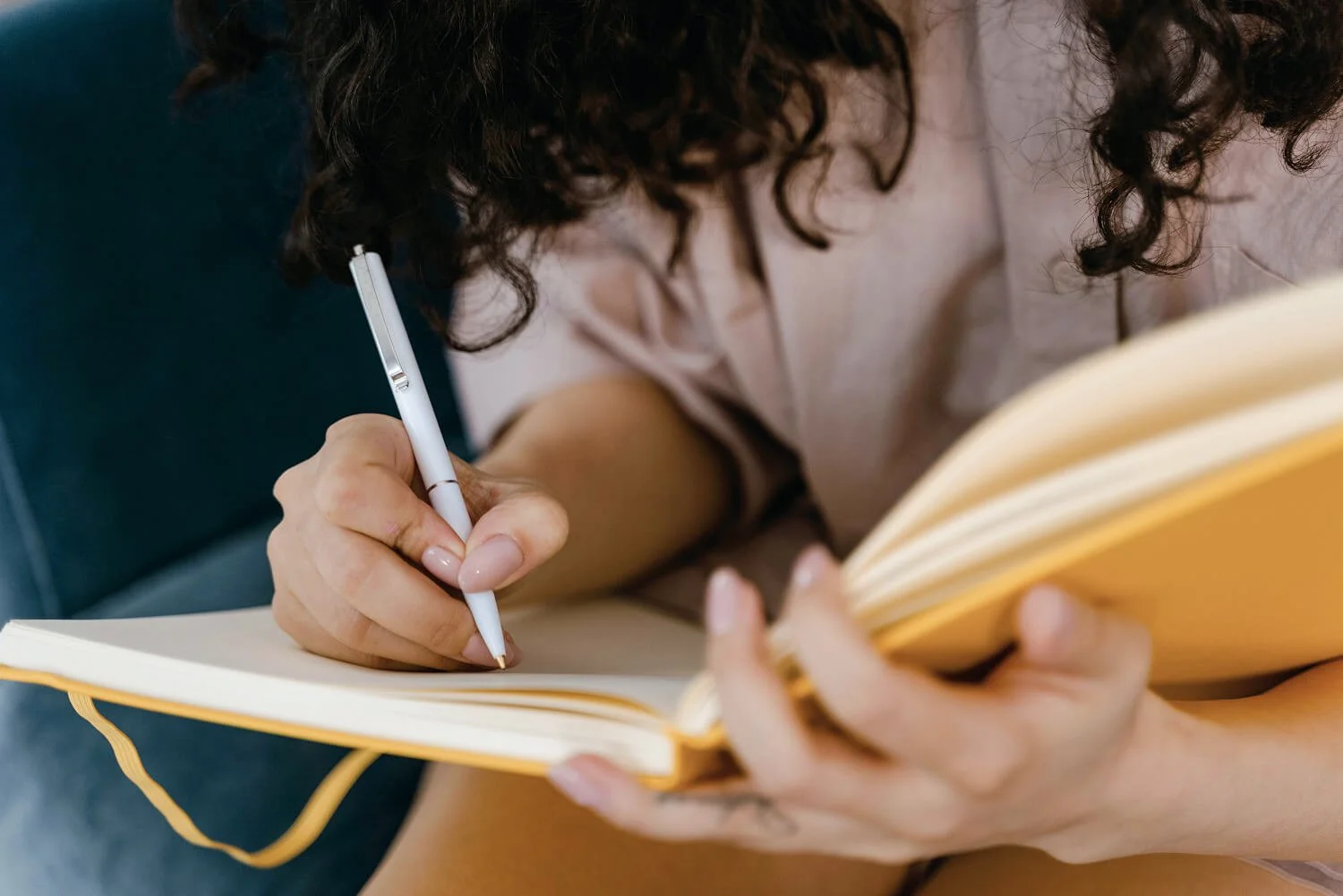 A woman with curly hair writing in a yellow notebook with a white pen.