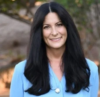 A woman with long black hair wearing a light blue top, smiling outdoors with trees in the background.