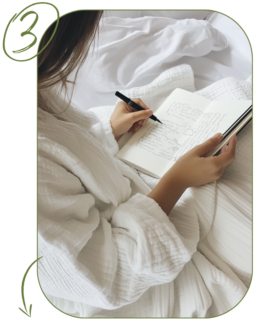 A woman in white pajamas sitting on a bed with white bedding, writing in a notebook with a black pen while holding a tablet.