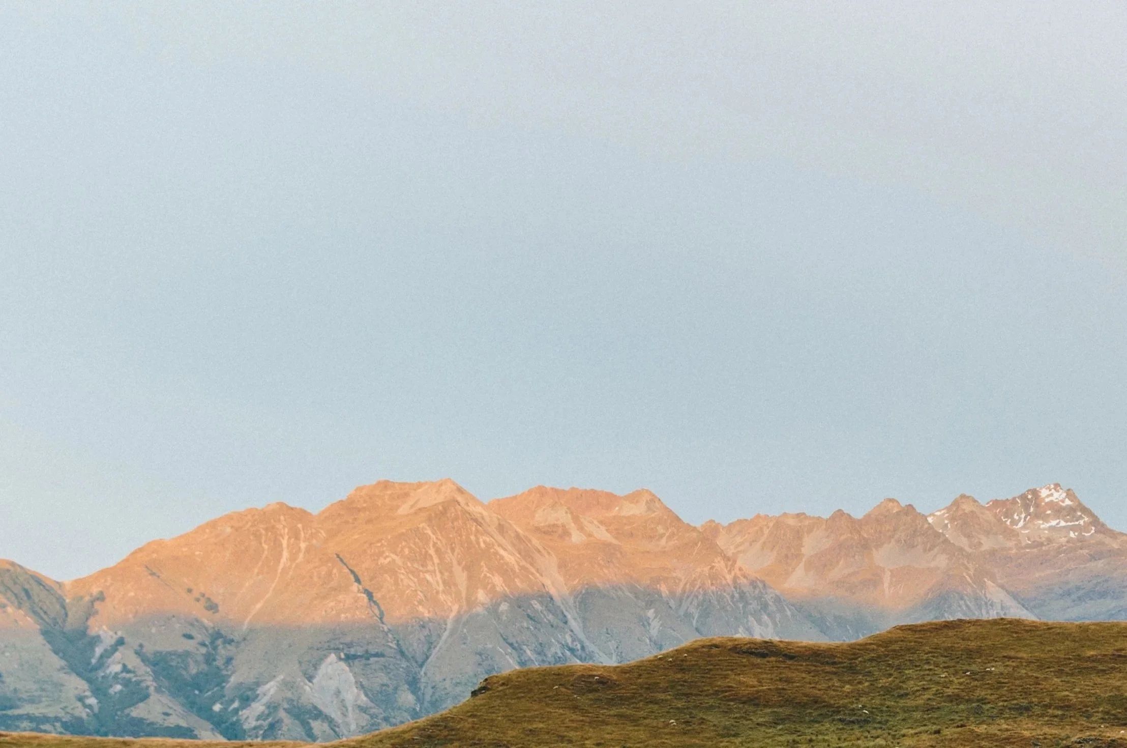 Cimes de montagnes avec sommet enneigé, en arrière-plan un ciel clair, au premier plan une colline verte.