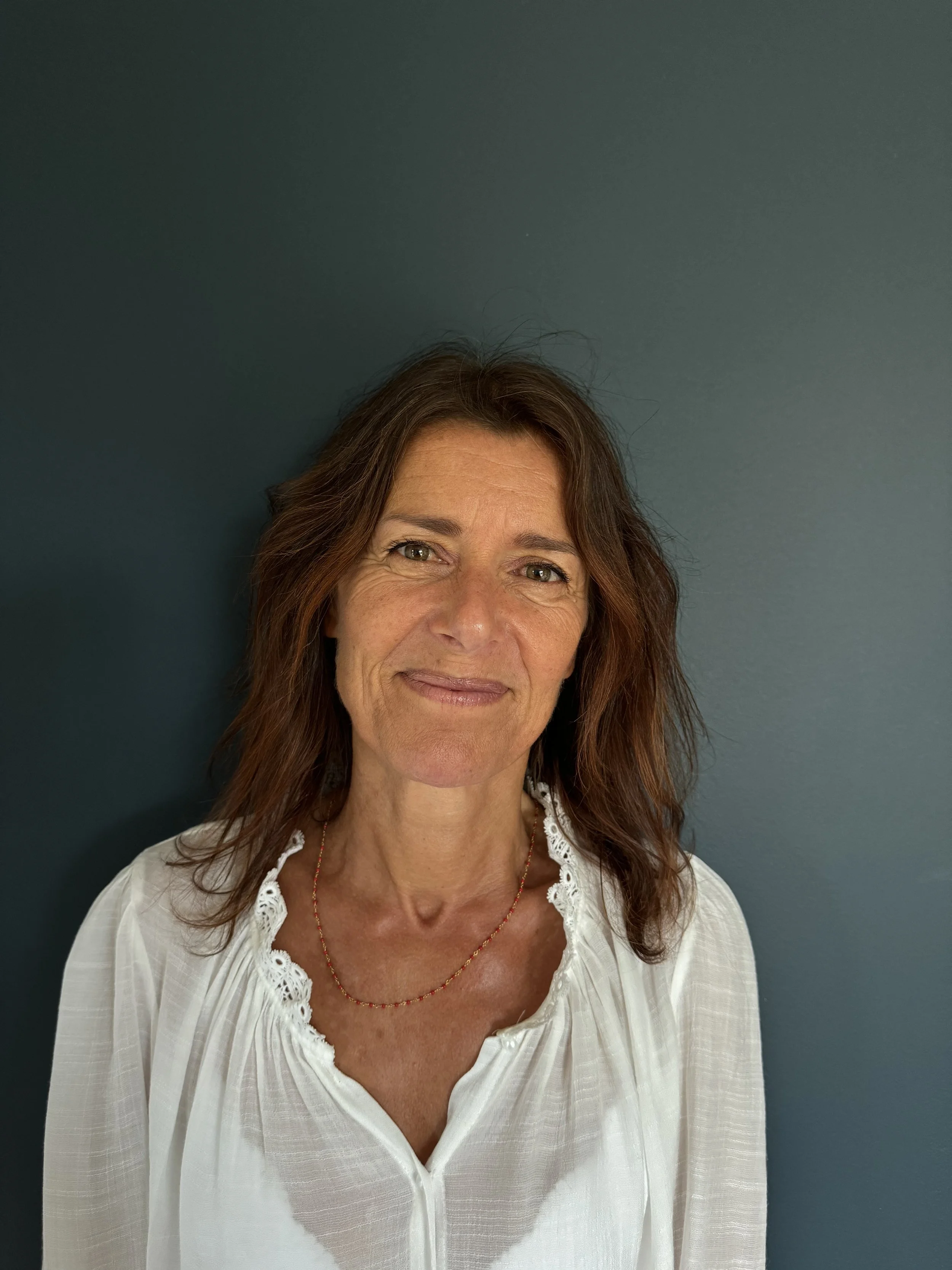 Portrait d'une femme aux cheveux bruns, portant un chemisier blanc et un collier, debout devant un mur bleu foncé.