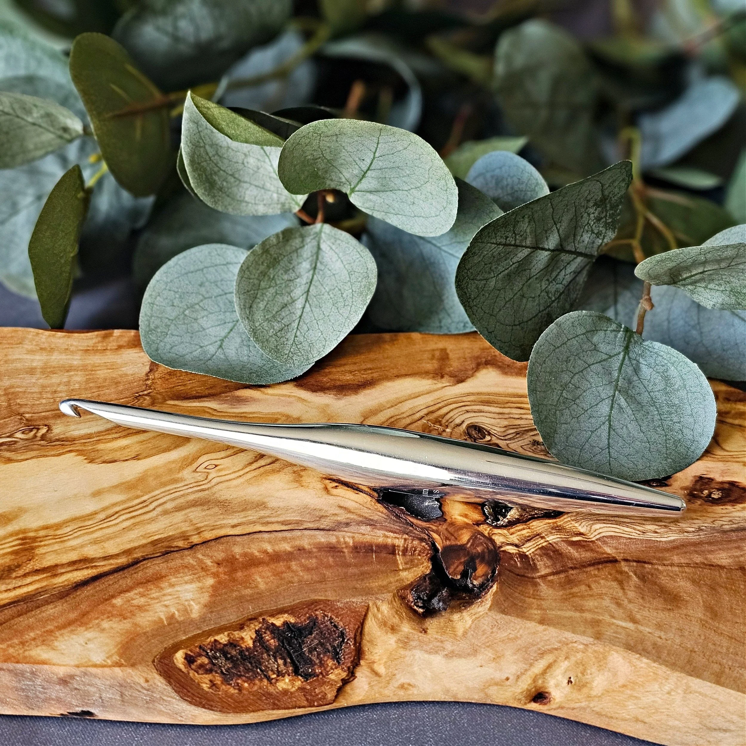 A silver metal letter opener resting on a piece of natural wood with green eucalyptus leaves in the background.