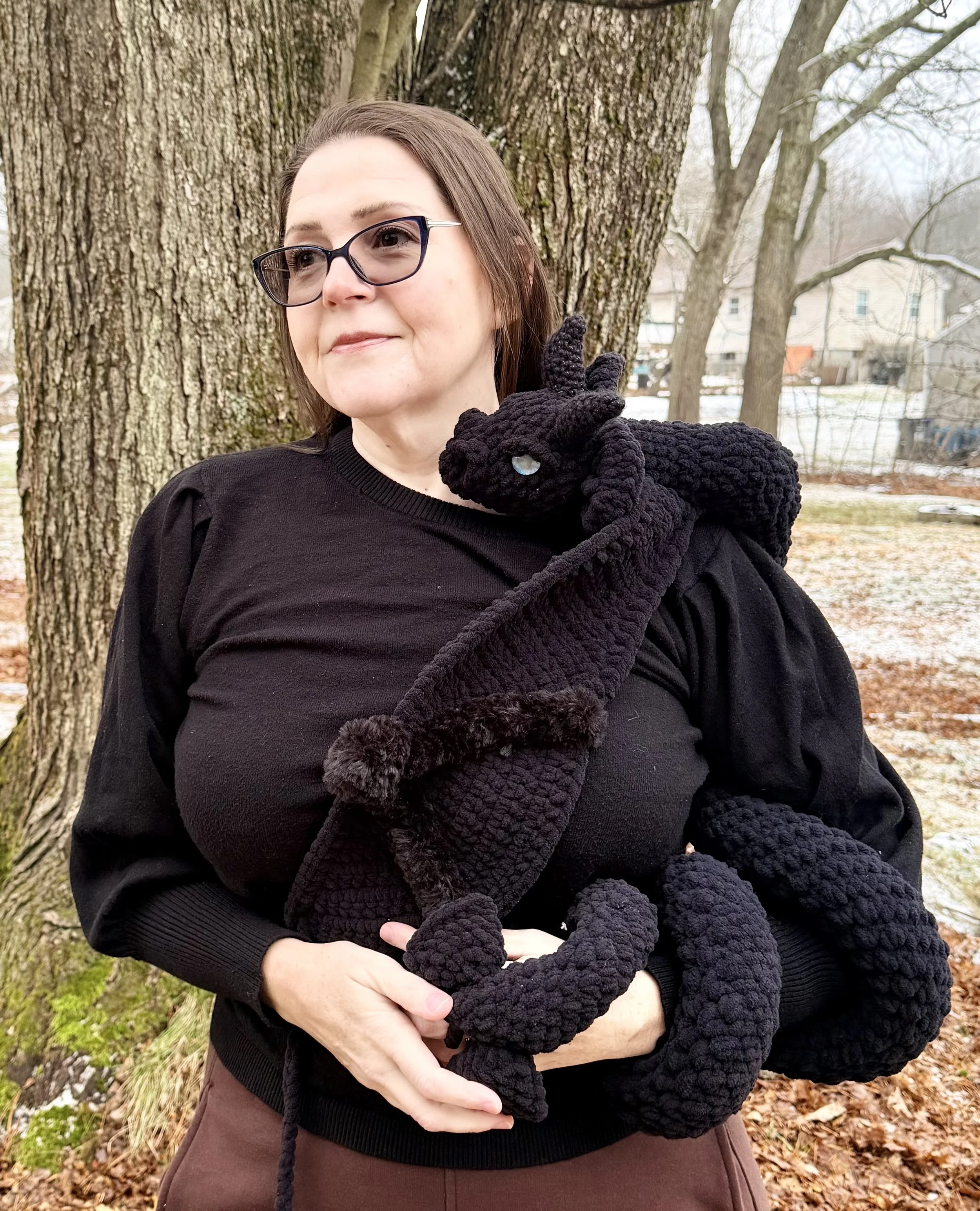A woman wearing glasses and a black sweater outdoors holding a large black crocheted dragon plush toy on her shoulder. There are trees and a residential neighborhood in the background.