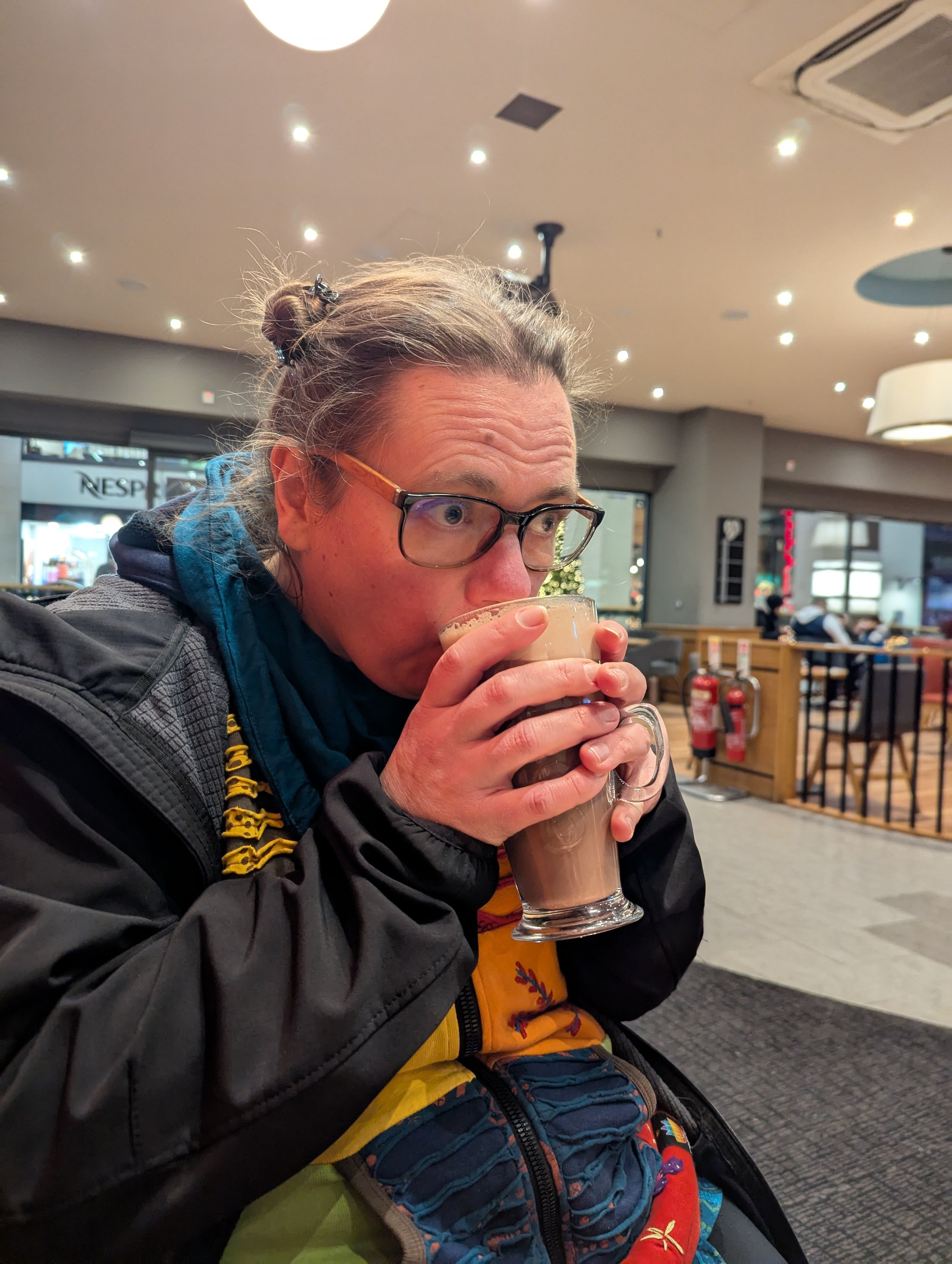 Ein Mensch mit Brille trinkt einen Kaffee oder Kakao in einem Café, sitzt an einem Tisch, im Hintergrund sind andere Gäste und eine verschneite Stadt draußen zu sehen.