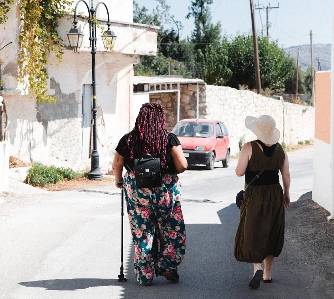Sabrina and her friend walk down the street in Greece, Sabrina uses her cane and wears bright floral pants, her friend is in a sun hat and a dark green jumper, their backs are turned away and they are deep in conversation.