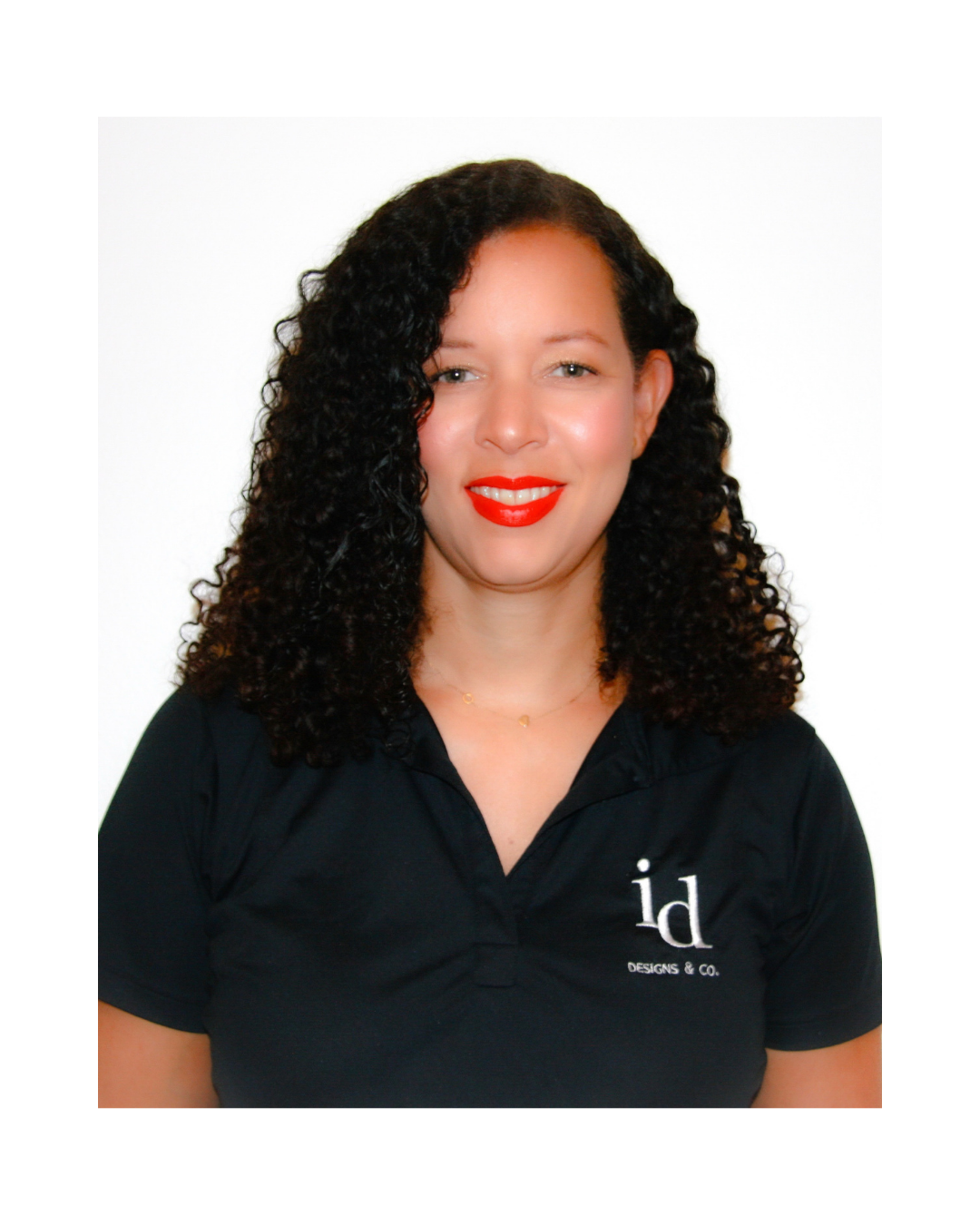 A woman with curly black hair, red lipstick, and a black collared shirt with an 'id Designs & Co.' logo, smiling at the camera against a plain background.
