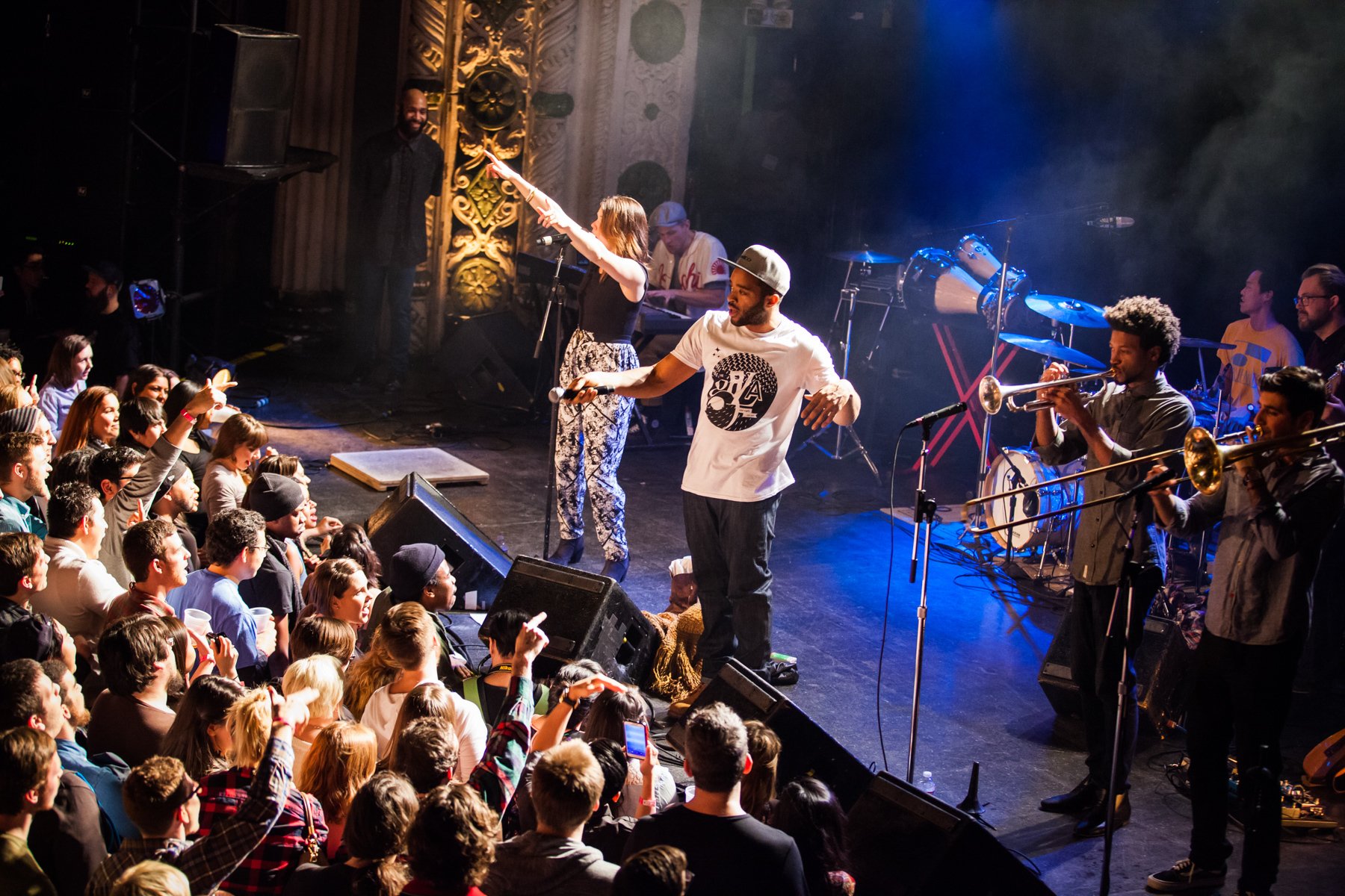 Musicians perform on stage with a singer, brass instruments, and percussion, while an audience watches and takes photos in a theater setting.