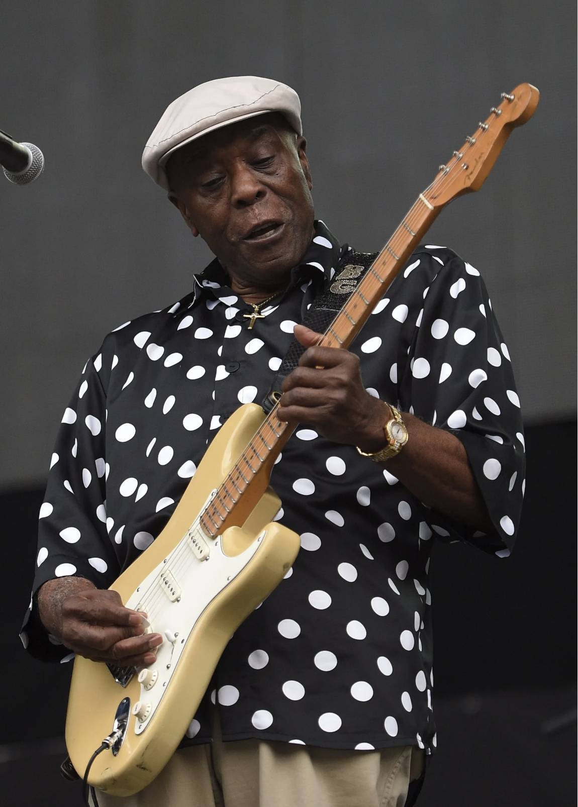 Buddy Guy playing a yellow electric guitar on stage, wearing a black and white polka dot shirt, beige pants, a white cap, a gold watch, and a gold necklace with a cross pendant.