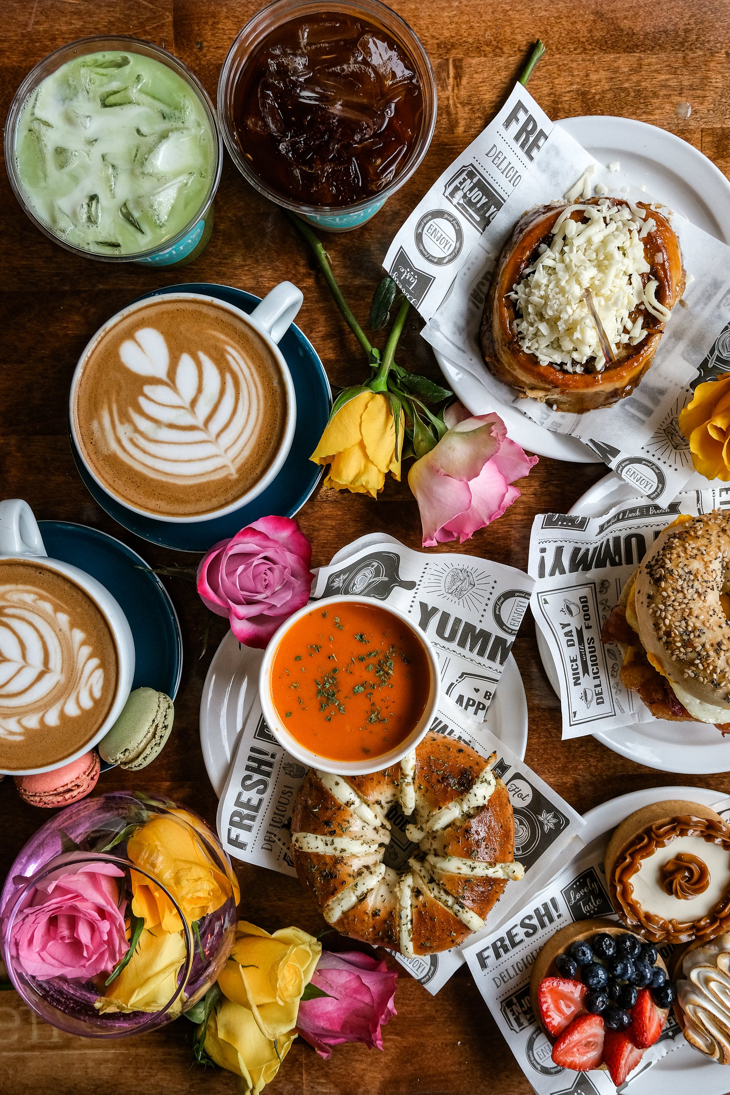 An assortment of drinks, desserts, and sandwiches on a wooden table, surrounded by colorful roses.