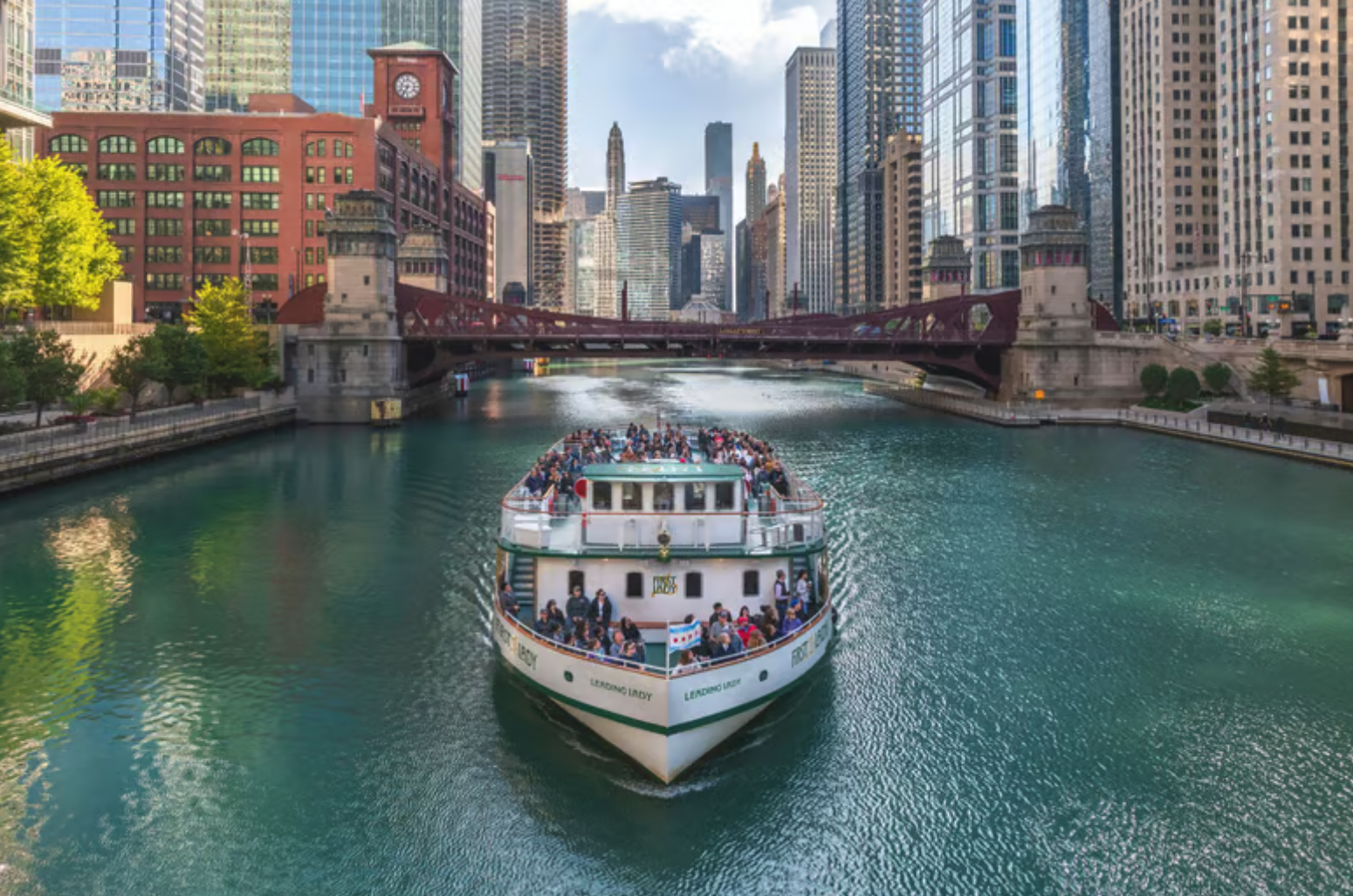 A river cruise boat with many passengers aboard traveling on a city river, surrounded by tall skyscrapers and buildings, with a bridge overhead.