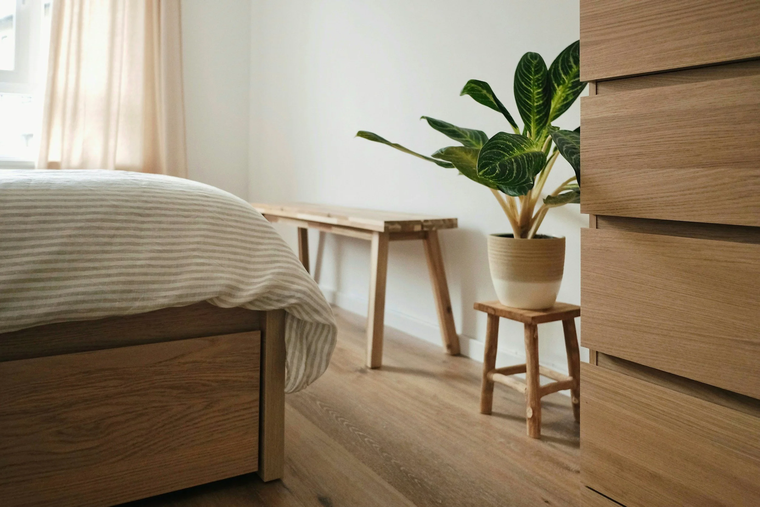 Ein Schlafzimmer mit einem Bett, einem kleinen Tisch aus Holz, einer Pflanze in einem Topf auf einem kleinen Holzstuhl, helle Wände und Vorhängen.