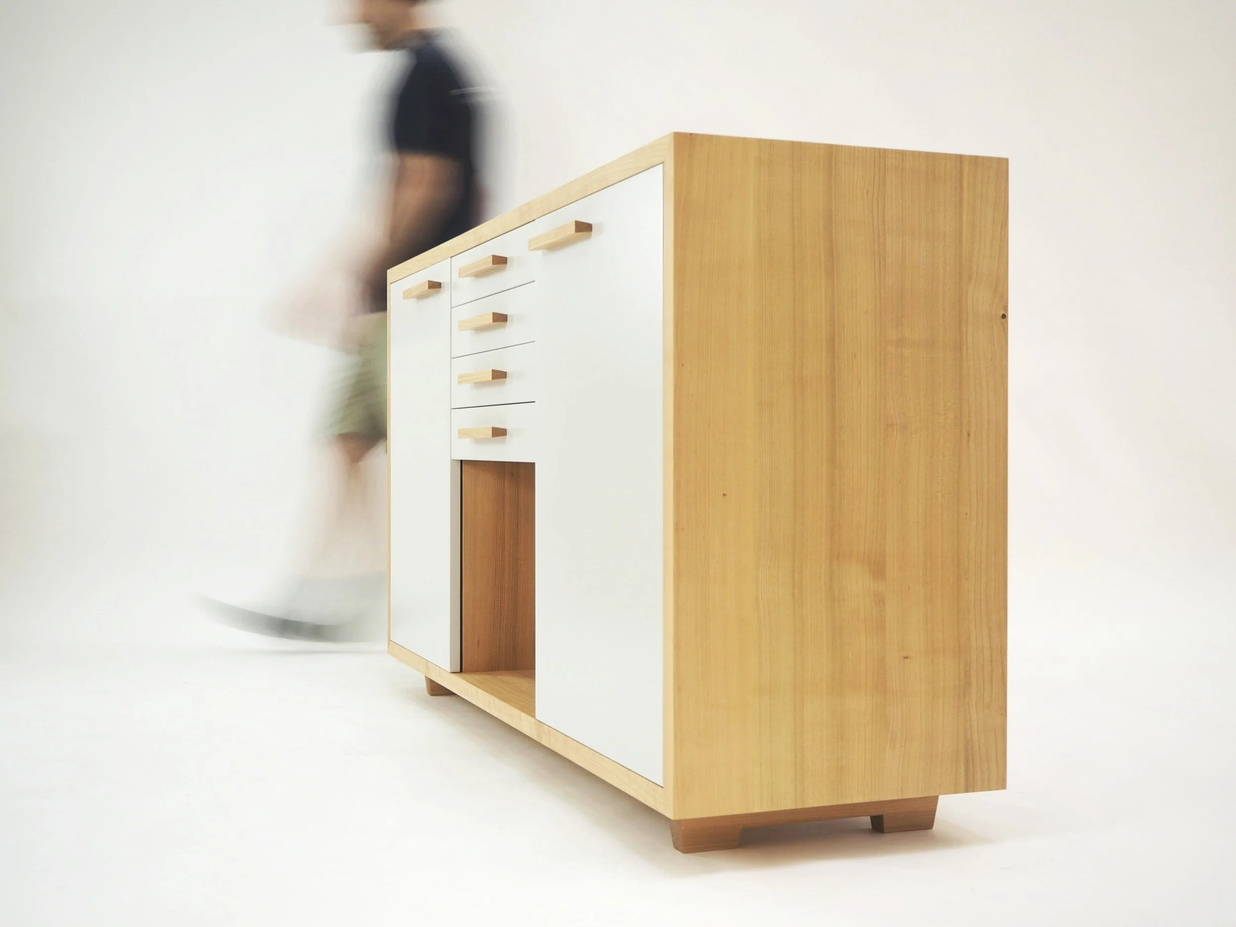 Moderner Holzschrank mit weißen Türen und Holzgriffen vor weißem Hintergrund, eine Person ist unscharf im Hintergrund.