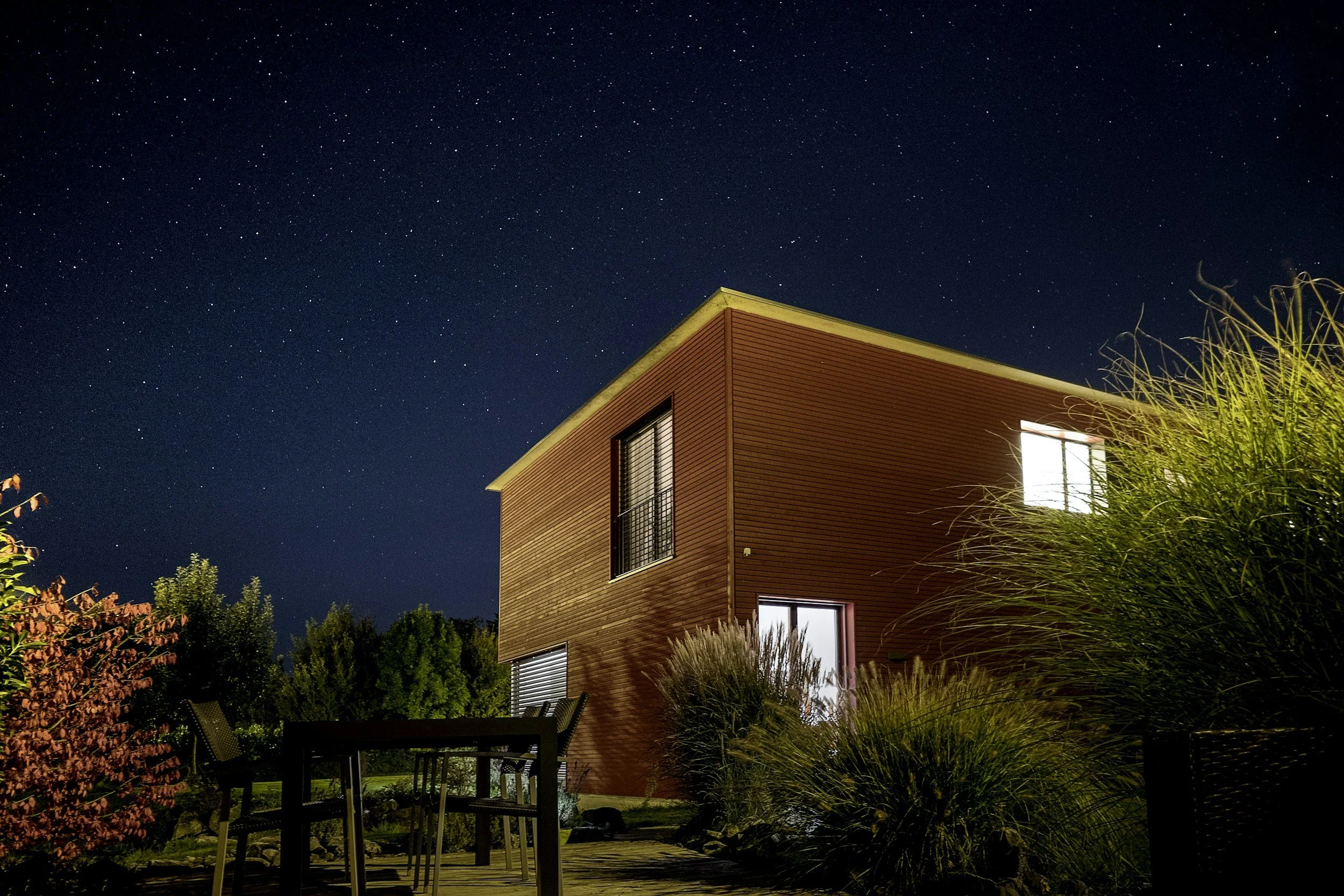 Modern zweistöckiges Haus bei Nacht mit sternenklarem Himmel im Hintergrund, beleuchtete Fenster, Garten mit Sitzgelegenheiten und großen Pflanzen im Vordergrund.