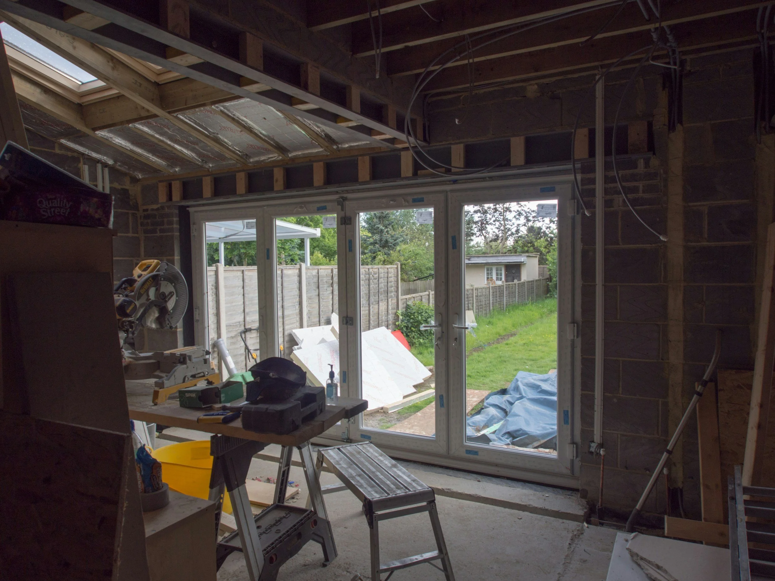 Innenansicht eines unfertigen Hauses mit großen Fenstern, Bauarbeiten und Werkzeugen, offenes Dach mit Dachisolierung, Garten im Hintergrund.